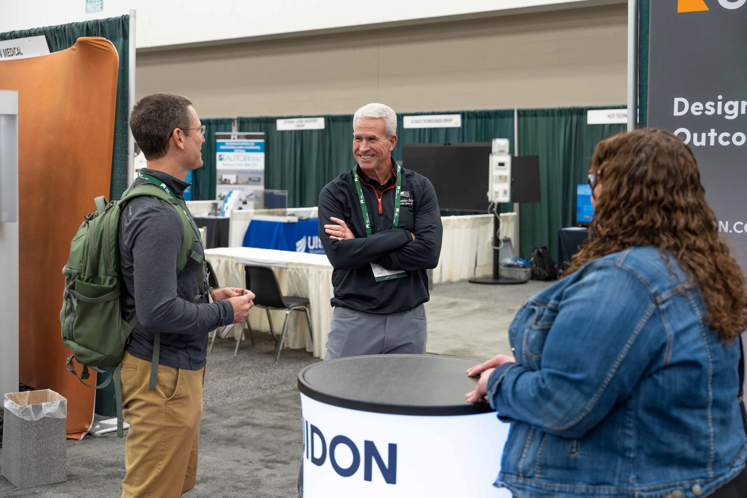 Three people are engaged in a conversation at an indoor event or trade show. A man with gray hair is smiling and standing with crossed arms, while a younger man with glasses and a backpack, and a woman with curly hair in a denim jacket listen. There 