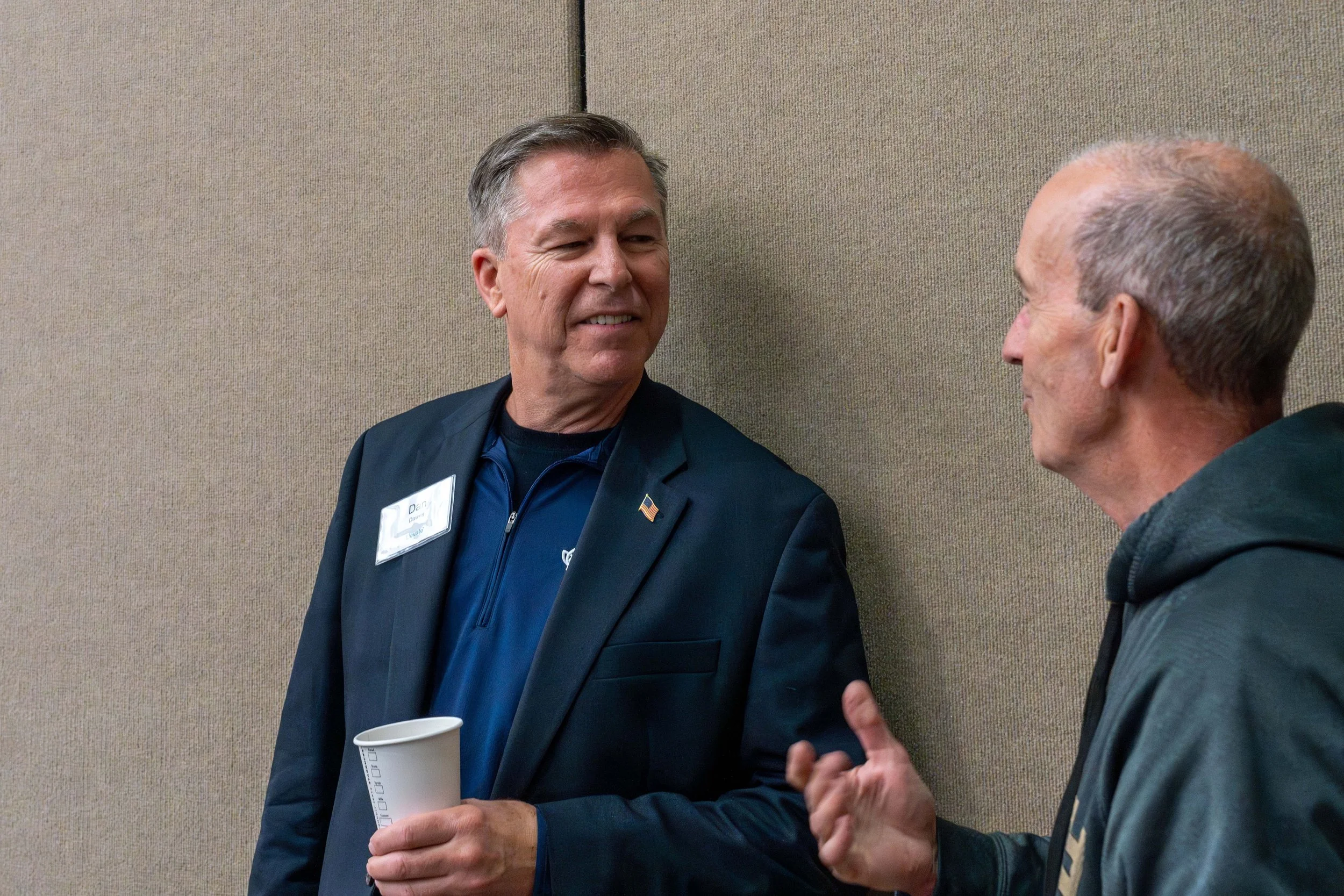 Two men are engaged in a conversation against a beige wall. One man is smiling and holding a paper cup, wearing a dark blazer with a name tag. The other man is gesturing with his hand, dressed in a hoodie.