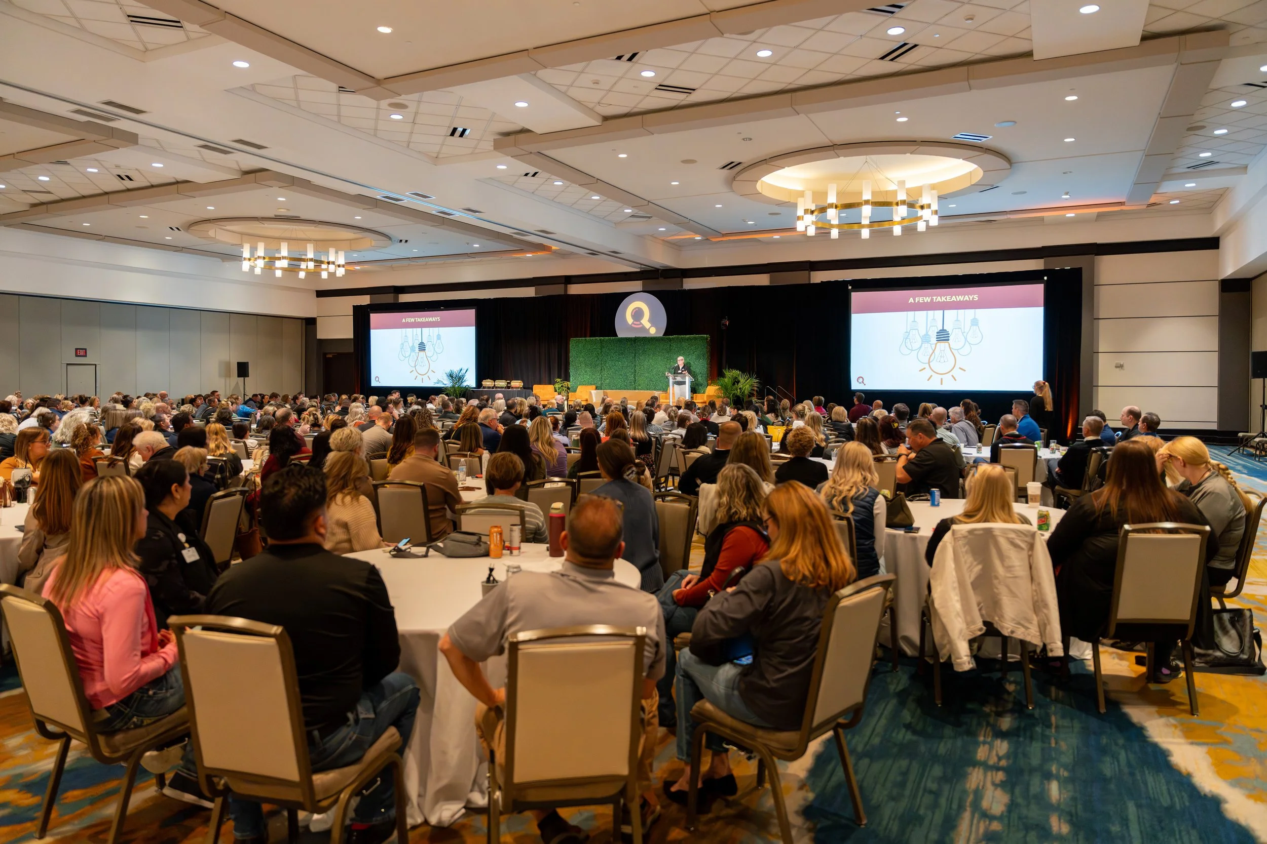 Large audience seated at round tables in a conference room, facing a stage with a speaker and two screens, during a presentation.