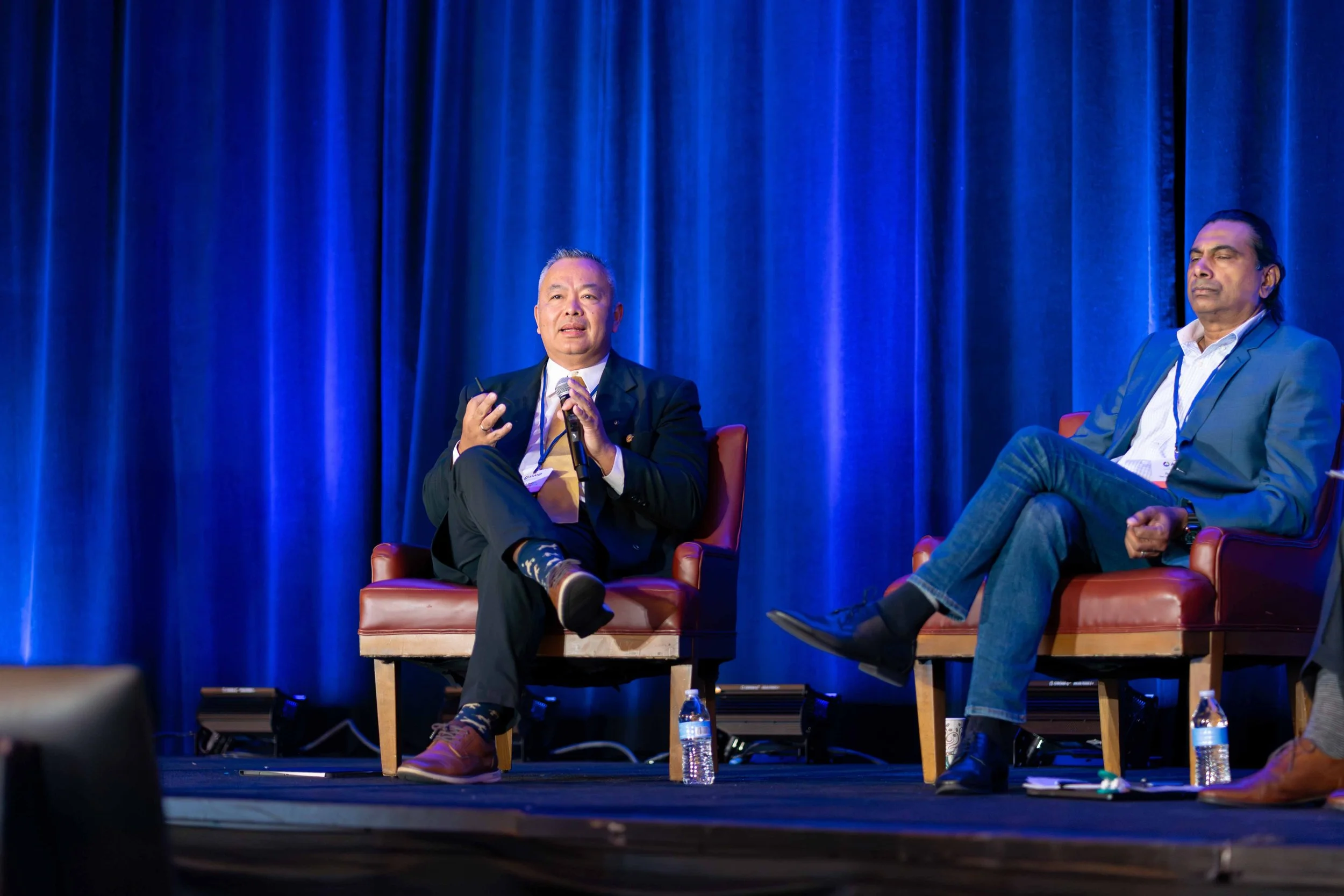 Two men sitting on stage chairs in front of a blue curtain, engaged in a conversation during a panel discussion or conference.