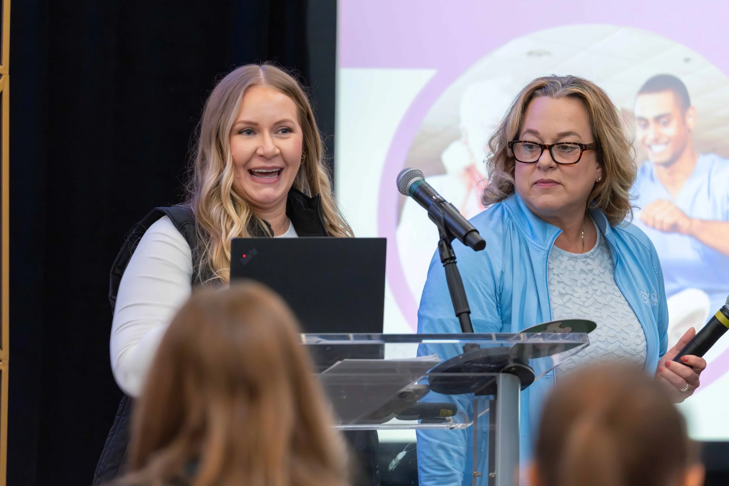 Two women standing behind a clear podium and speaking into a microphone at a conference. The woman on the left has long blonde hair, is smiling, and wearing a white top with a black vest. The woman on the right has shoulder-length blonde hair, glasse