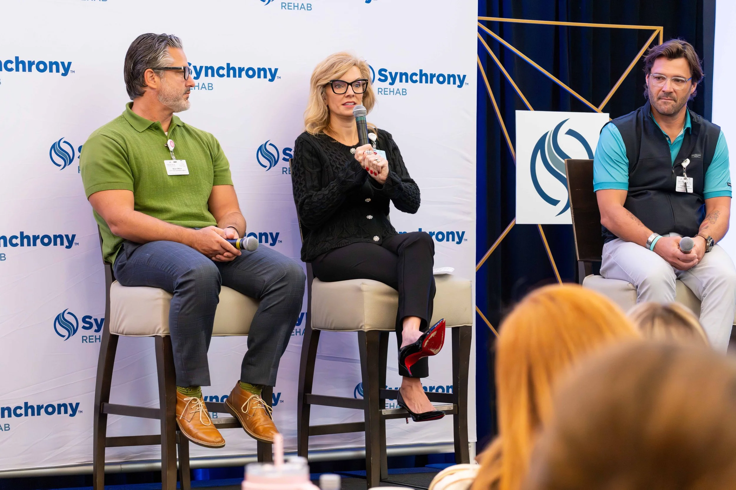 Three individuals seated on stage during a panel discussion at Synchrony Rehab event, with a woman speaking into a microphone between two men, all wearing name badges, in front of a backdrop displaying the Synchrony logo.