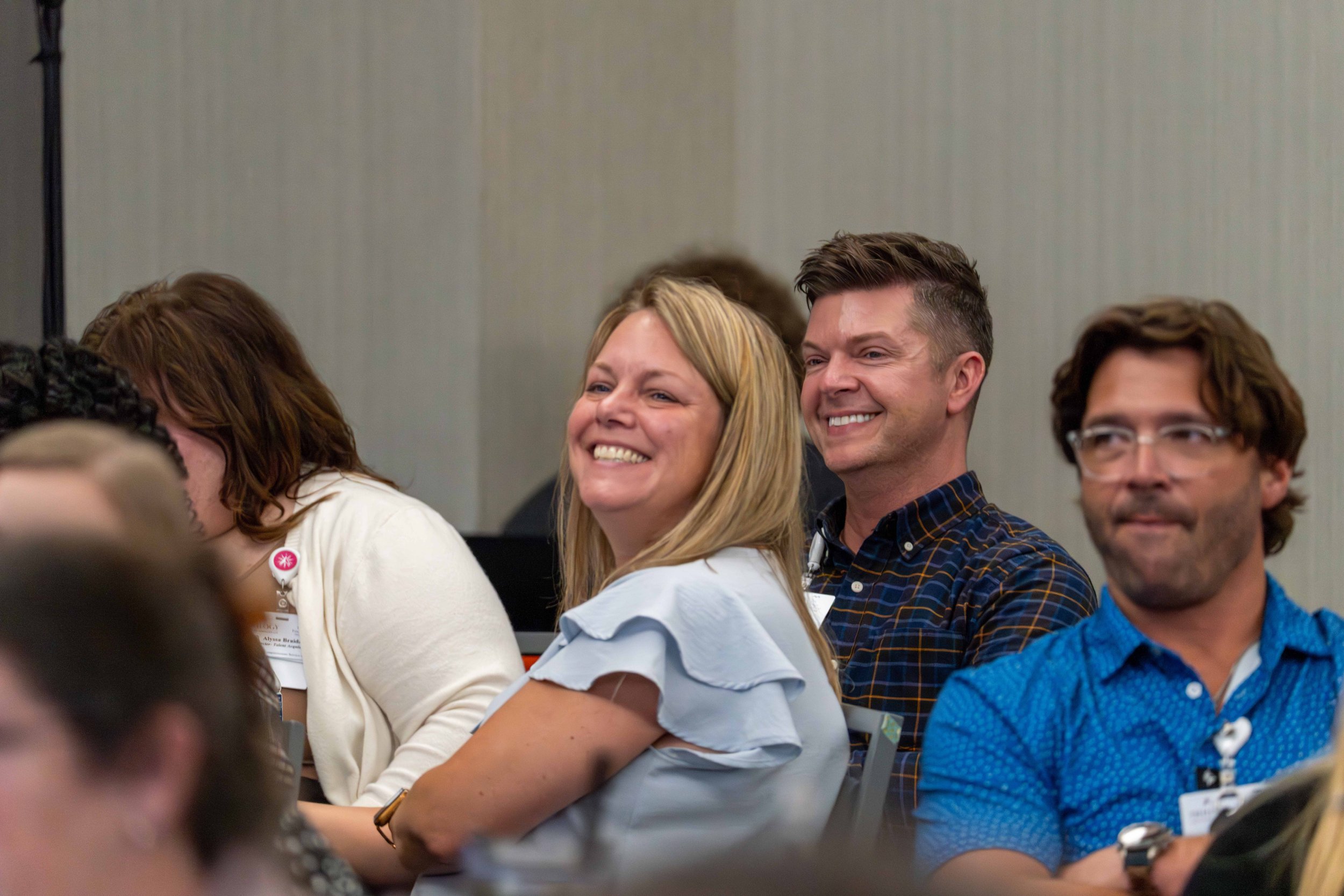 A group of people sitting and smiling at an indoor event, with a woman in a light blue blouse and a man in a checked shirt in focus.
