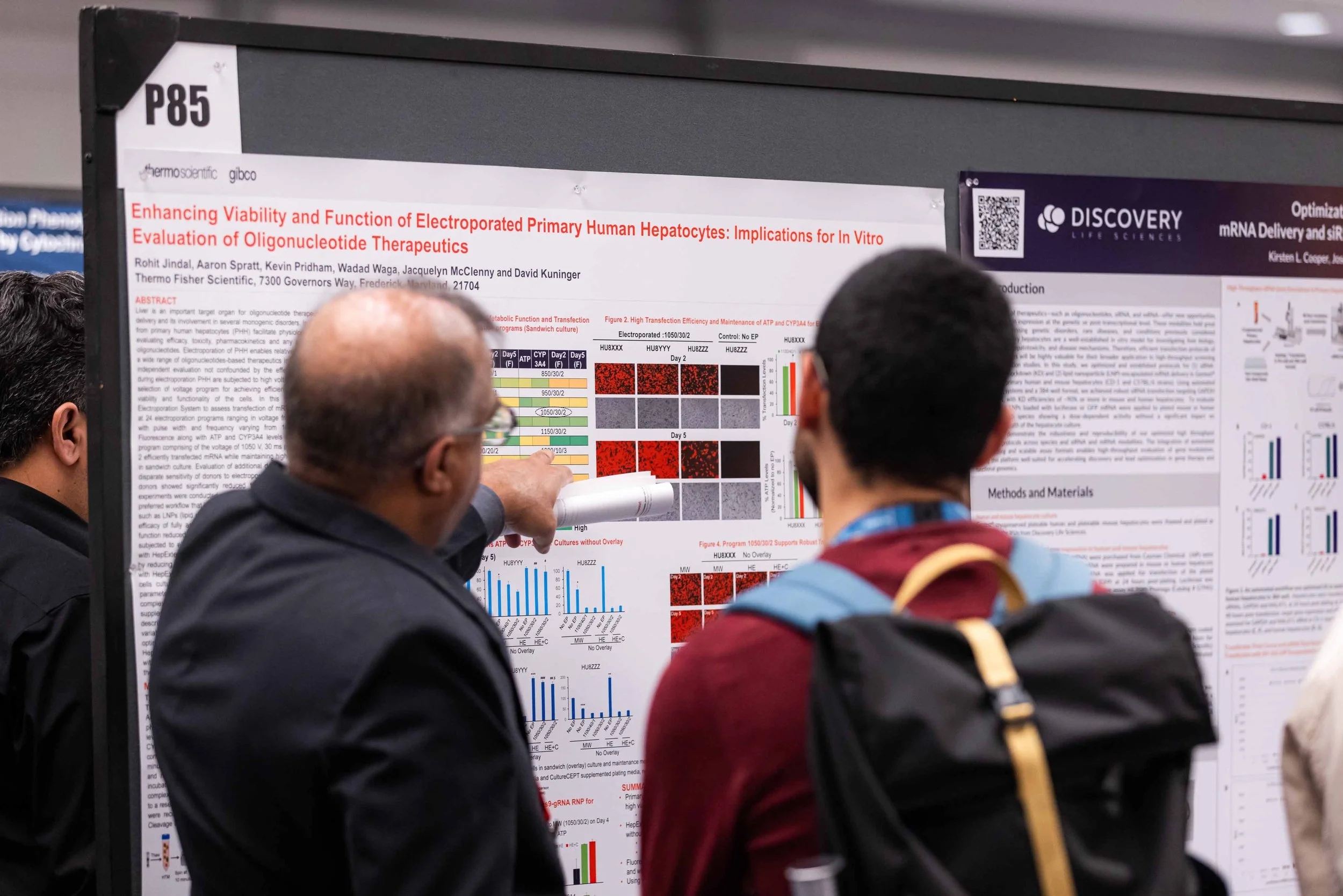 Two men are discussing a scientific research poster at a conference. The poster includes graphs, charts, and text about hepatocyte research.