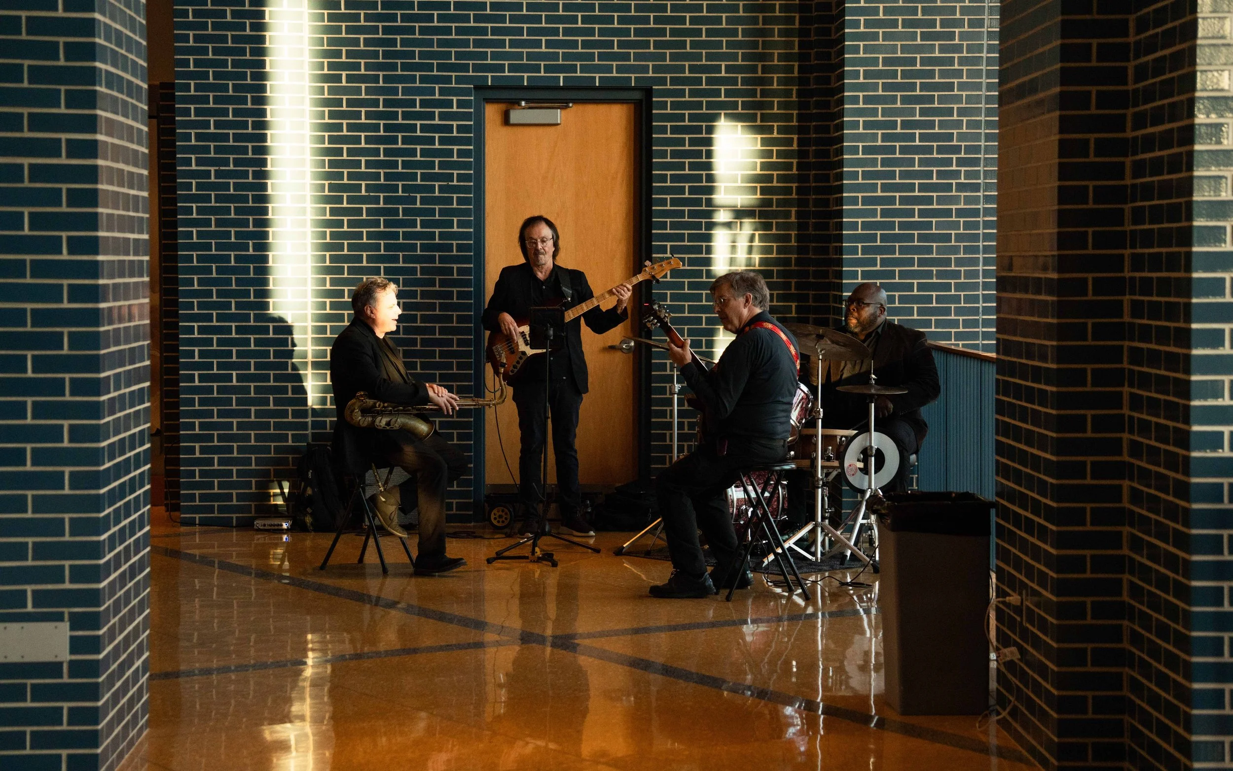 Four musicians playing jazz instruments in an indoor venue with blue brick walls and wooden flooring.