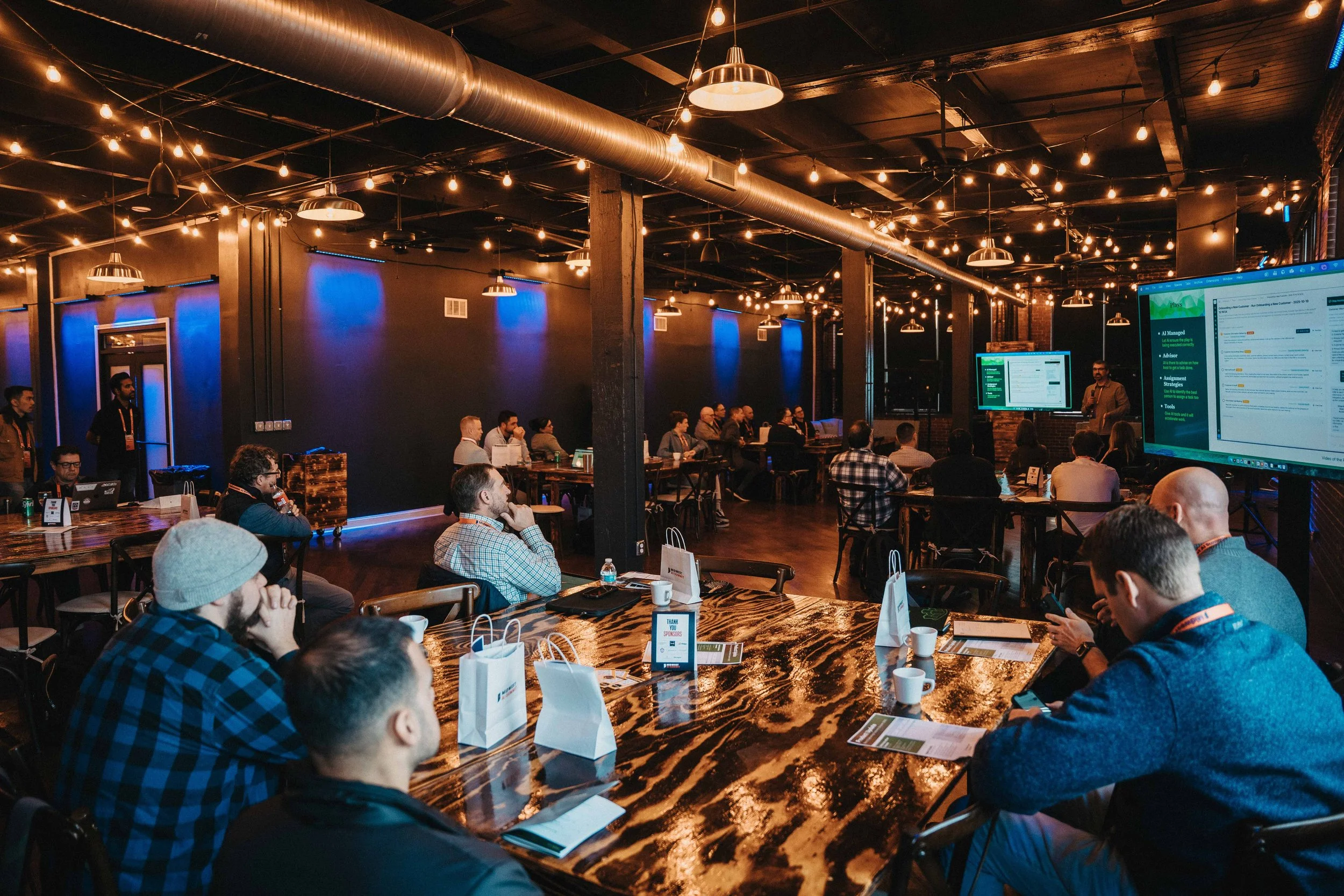 A group of people attending a presentation or conference in a dimly lit venue with string lights and modern decor, sitting around tables with laptops, papers, and coffee cups, while a speaker presents slides on large screens at the front.