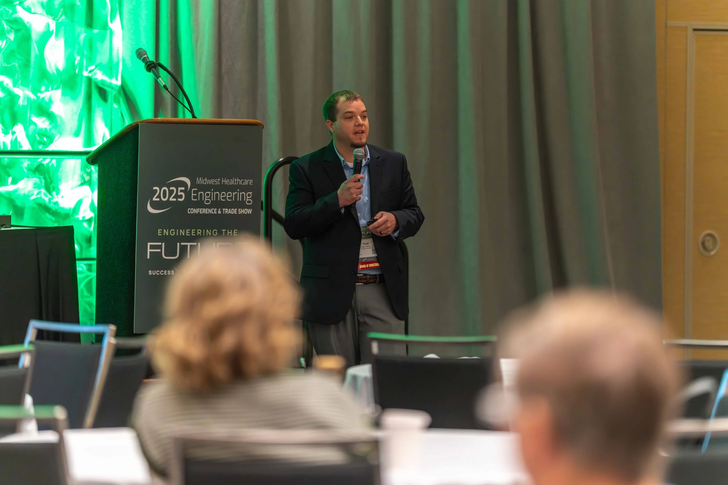 A man giving a presentation at a conference, standing beside a podium with a microphone, in front of an audience, with a green-lit backdrop. The podium displays text related to a healthcare engineering conference.