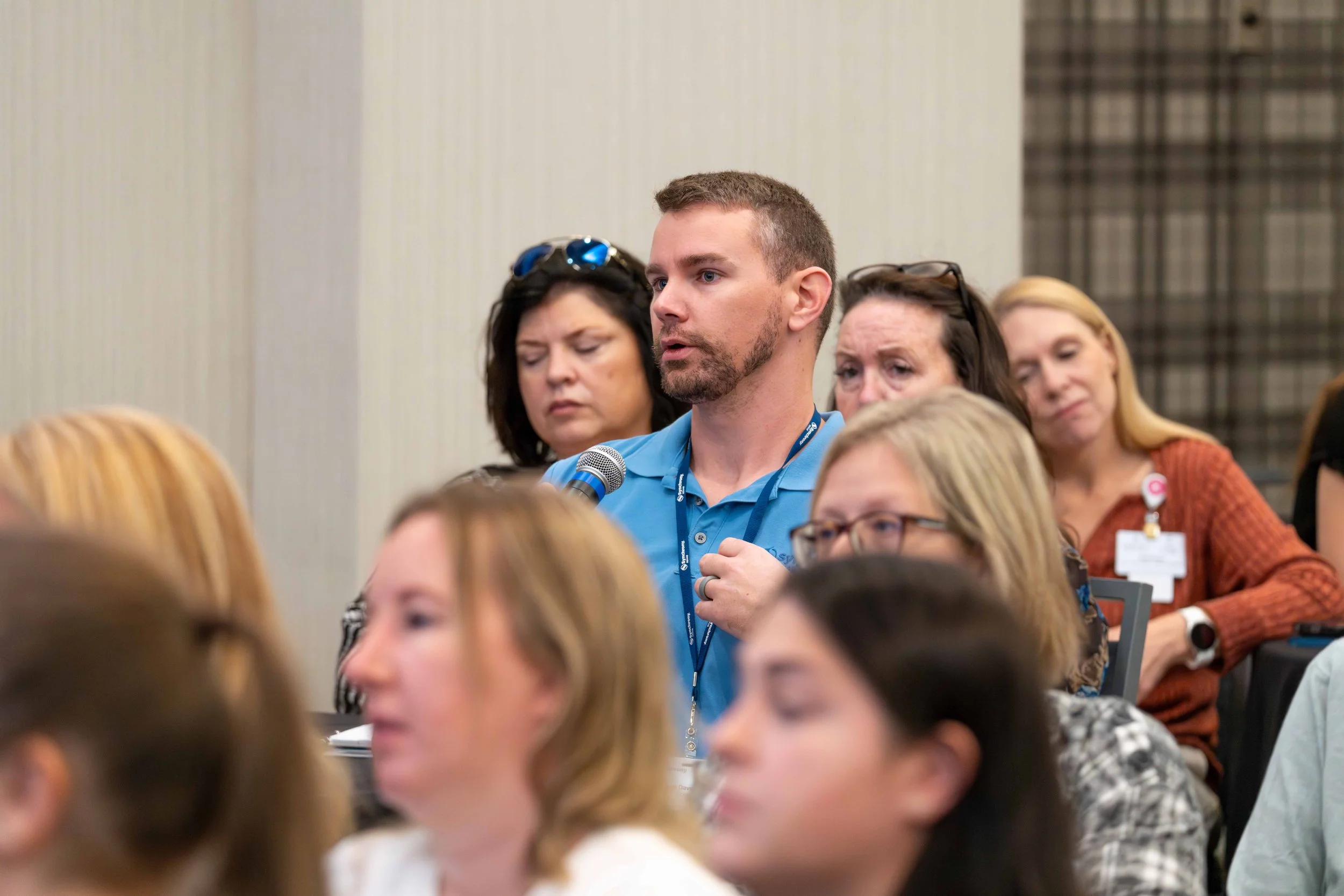 A man in a blue shirt holding a microphone appears to be speaking during a conference or seminar, with several women seated around him listening attentively.