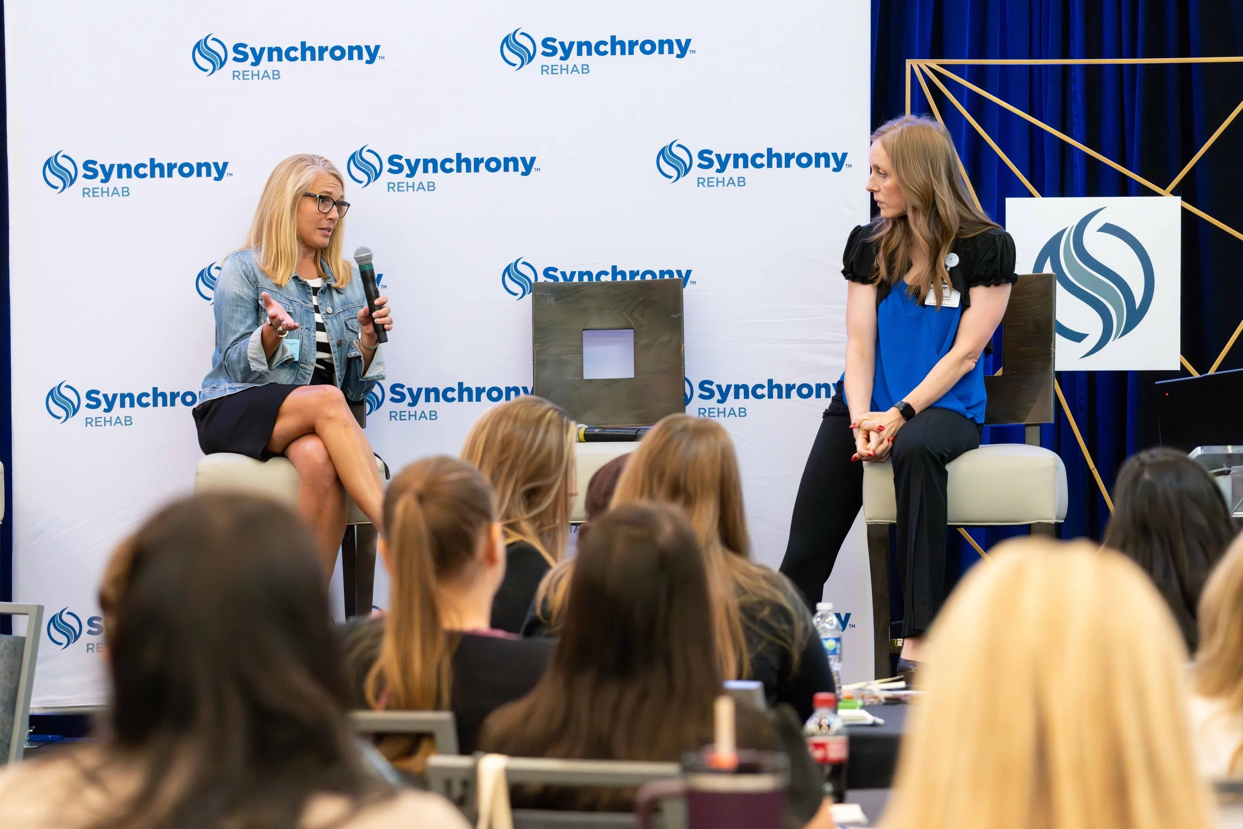 Two women on stage having a discussion at an event with a backdrop displaying the Synchrony Rehab logo. One woman, blonde with glasses, is speaking into a microphone, while the other woman with long brunette hair is listening. Audience members are se