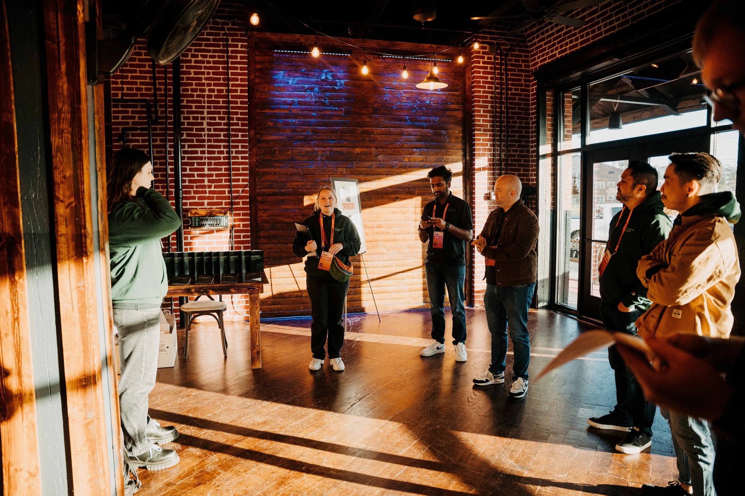 Group of people gathered inside a brick-walled venue for a presentation or tour, with sunlight streaming through large windows on a bright day.