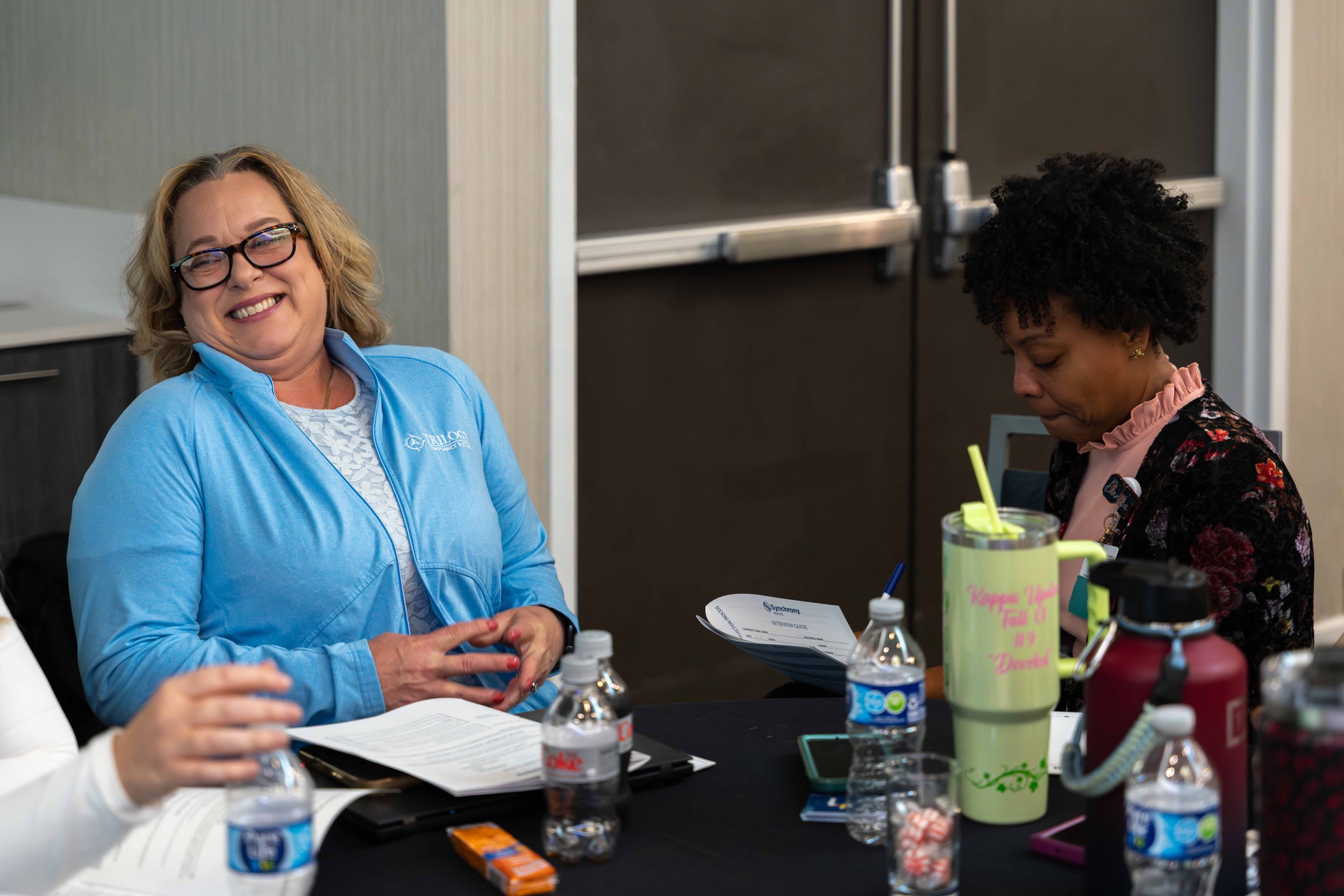 Two women sitting at a conference table, smiling and engaged in a discussion, surrounded by water bottles, papers, and colorful drink containers.