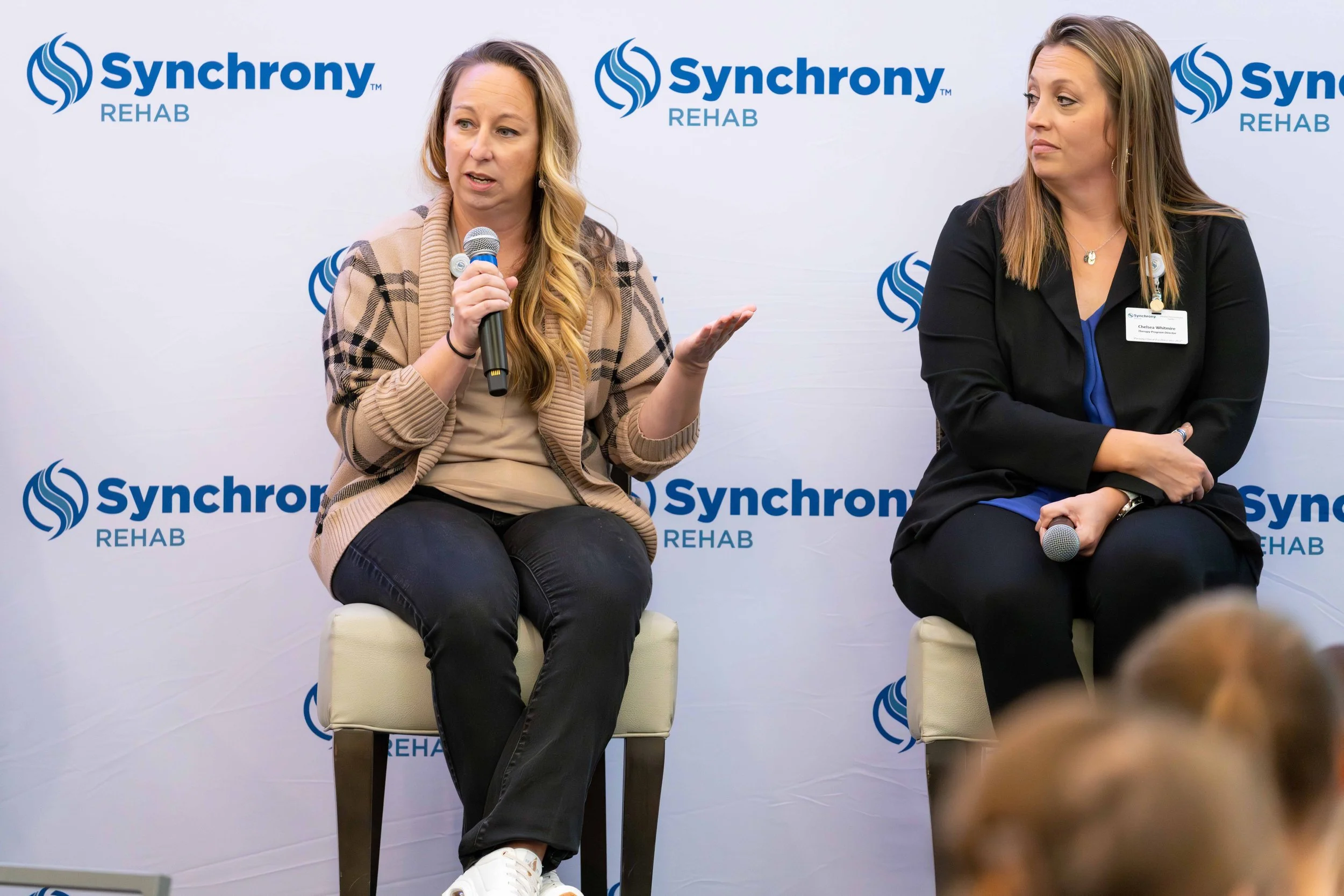 Two women in a panel discussion at Synchrony Rehab event. The woman on the left is speaking into a microphone, wearing a beige and black plaid cardigan and black pants. The woman on the right is sitting with a microphone in her hand, wearing a black 