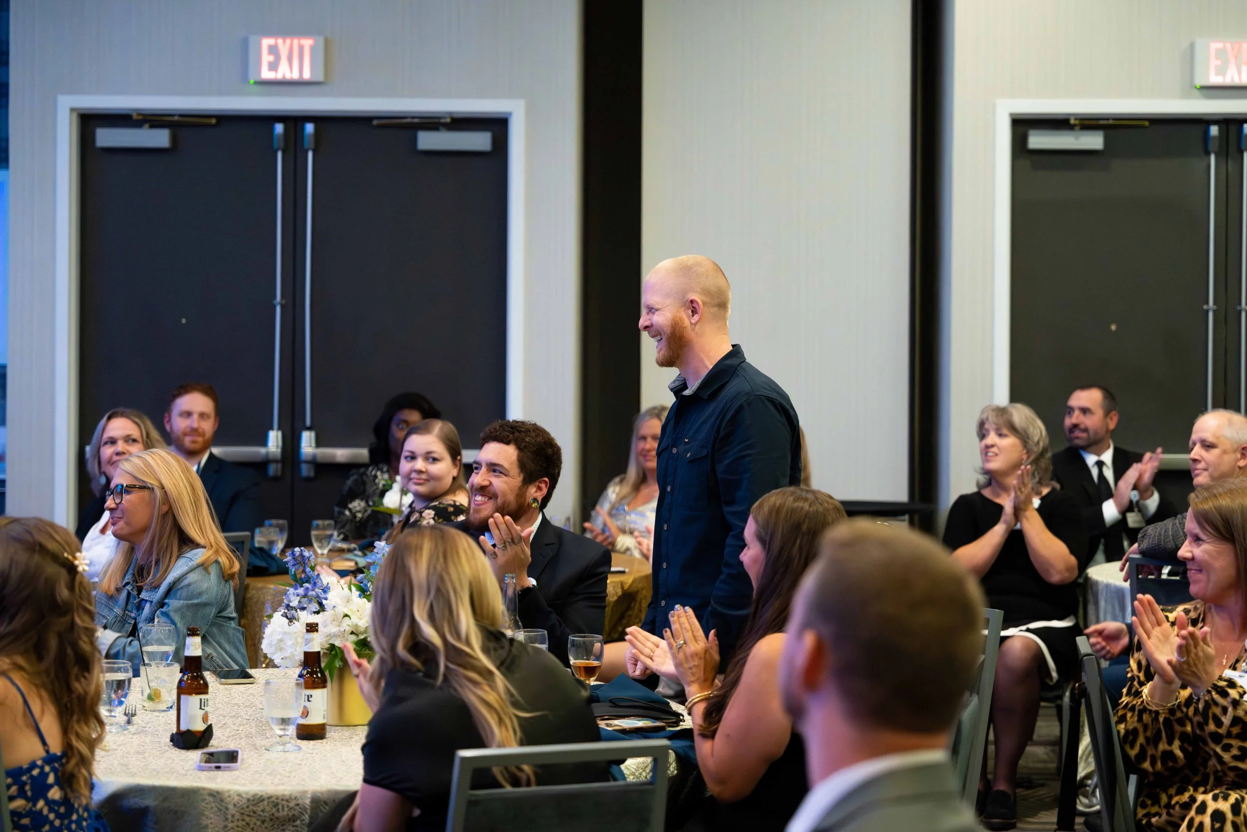 A man standing and smiling at a formal dinner or celebration event surrounded by seated guests who are clapping and looking at him, indoors with black exit doors in the background.