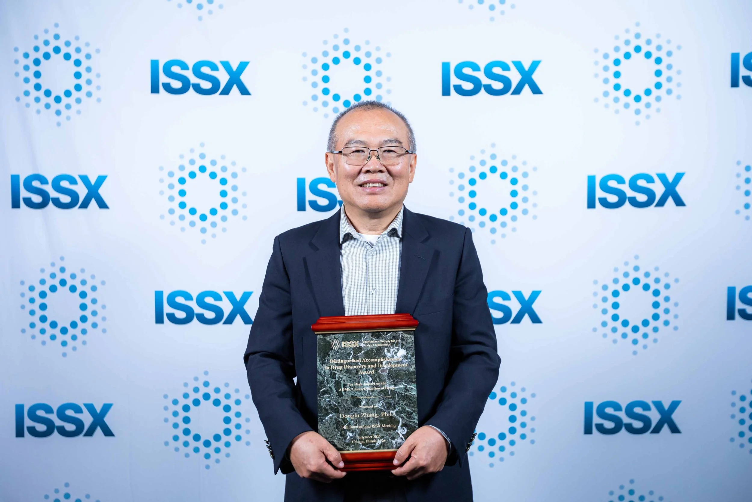 A man in a suit holding an award plaque, standing in front of a white background with the ISSX logo repeated in blue.