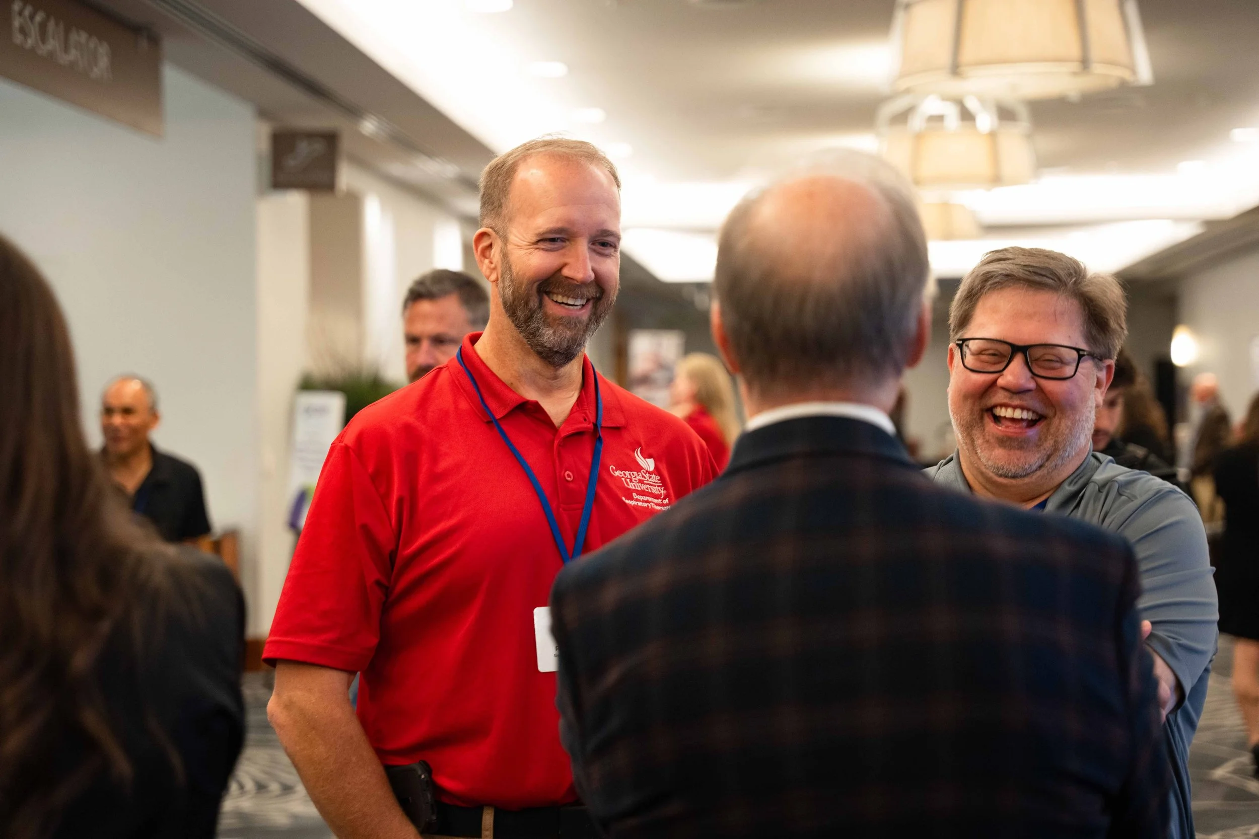 People smiling and talking at an indoor event or conference, with a man in a red shirt, a man in glasses, and a man with his back turned