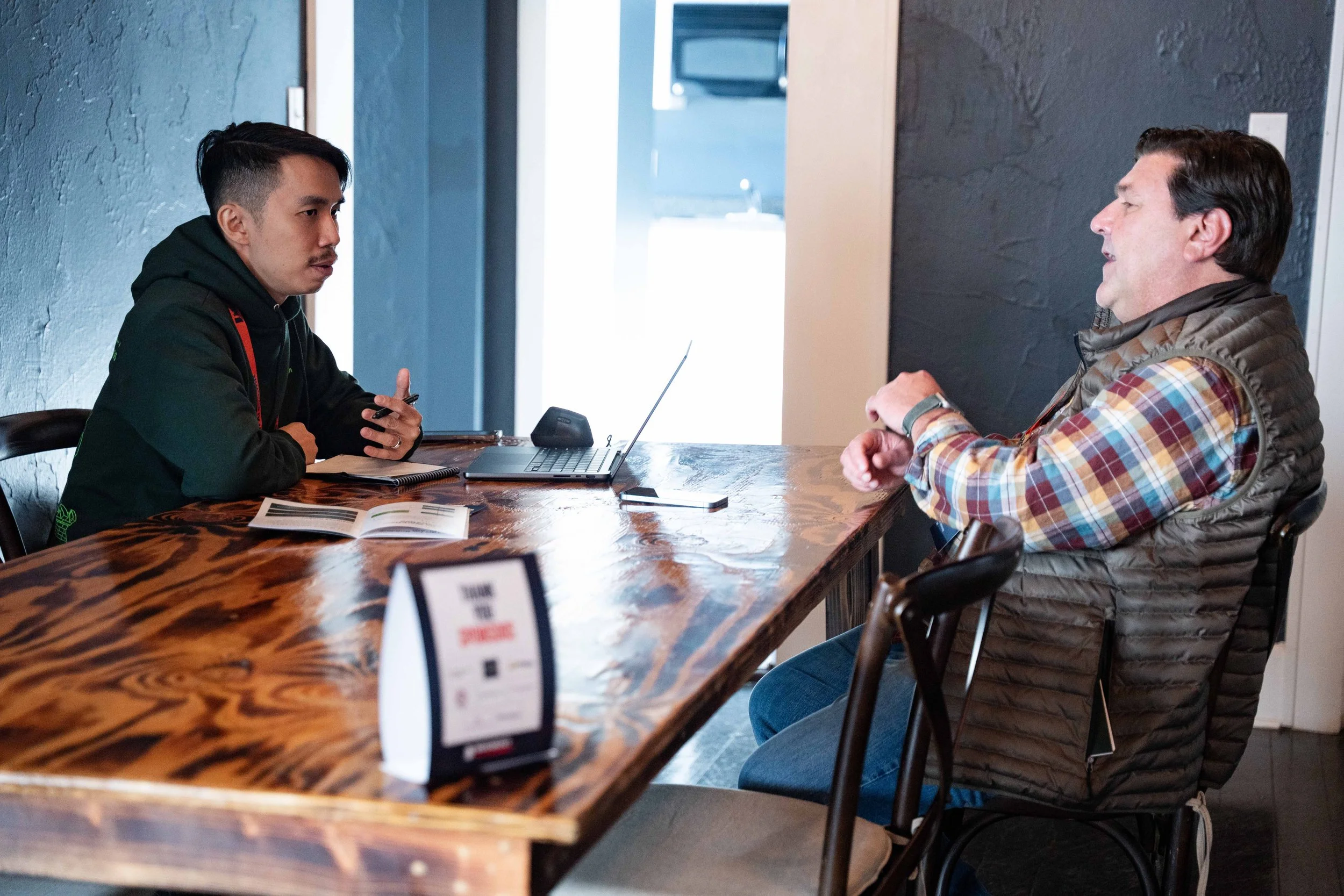 Two men sitting at a wooden table having a conversation in a modern casual setting. One man appears to be explaining something while the other listens attentively.