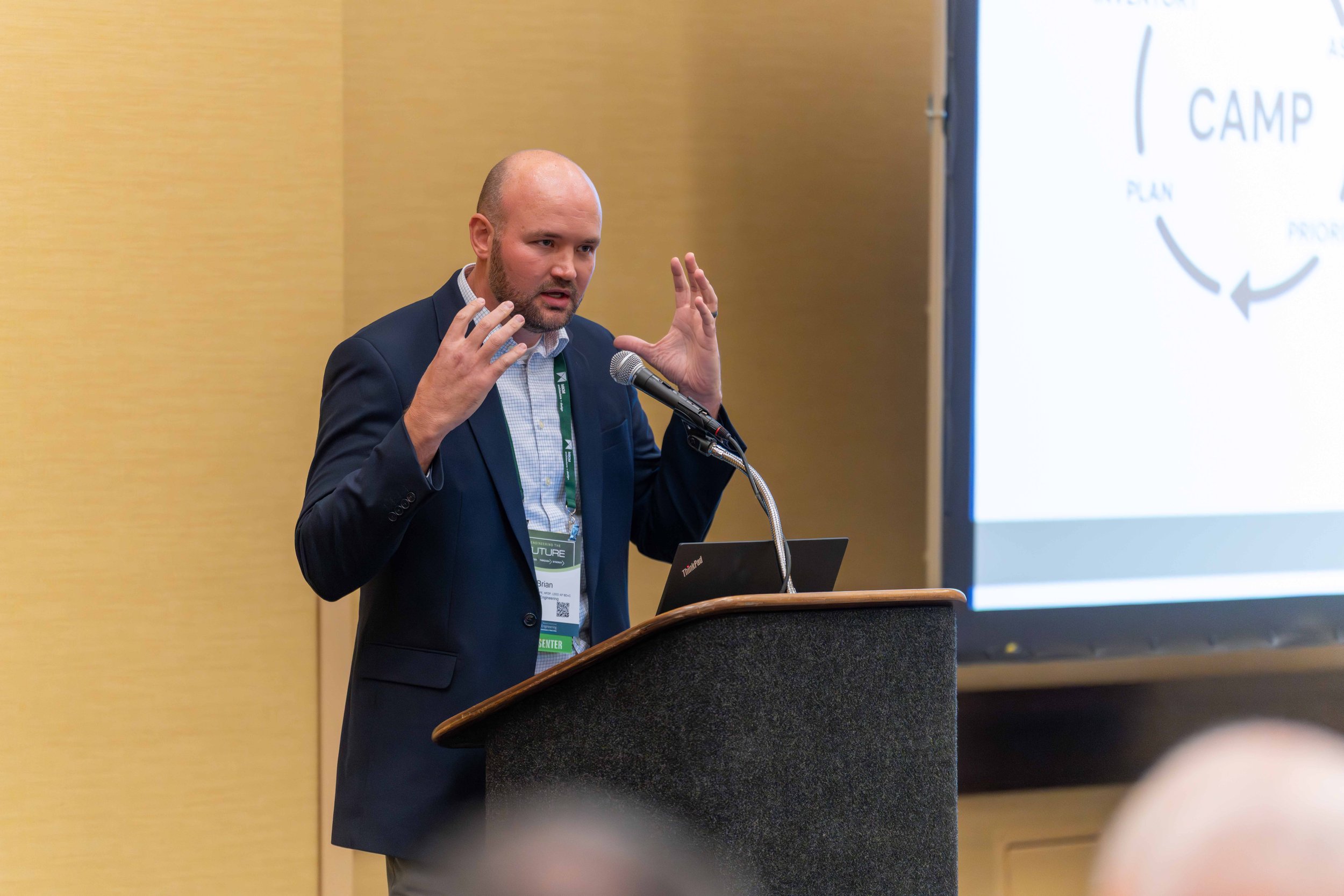 A man in a dark suit and light blue shirt is speaking at a podium with a microphone, gesturing with his hands, during a presentation at a conference.