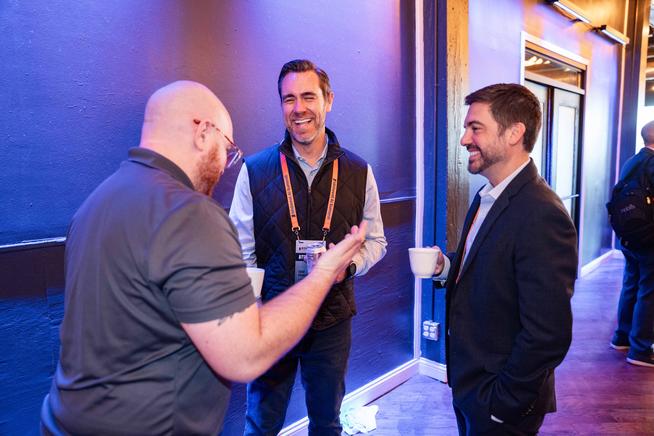 Three men smiling and talking at a social event, holding cups, in a room with purple walls and wooden floors.