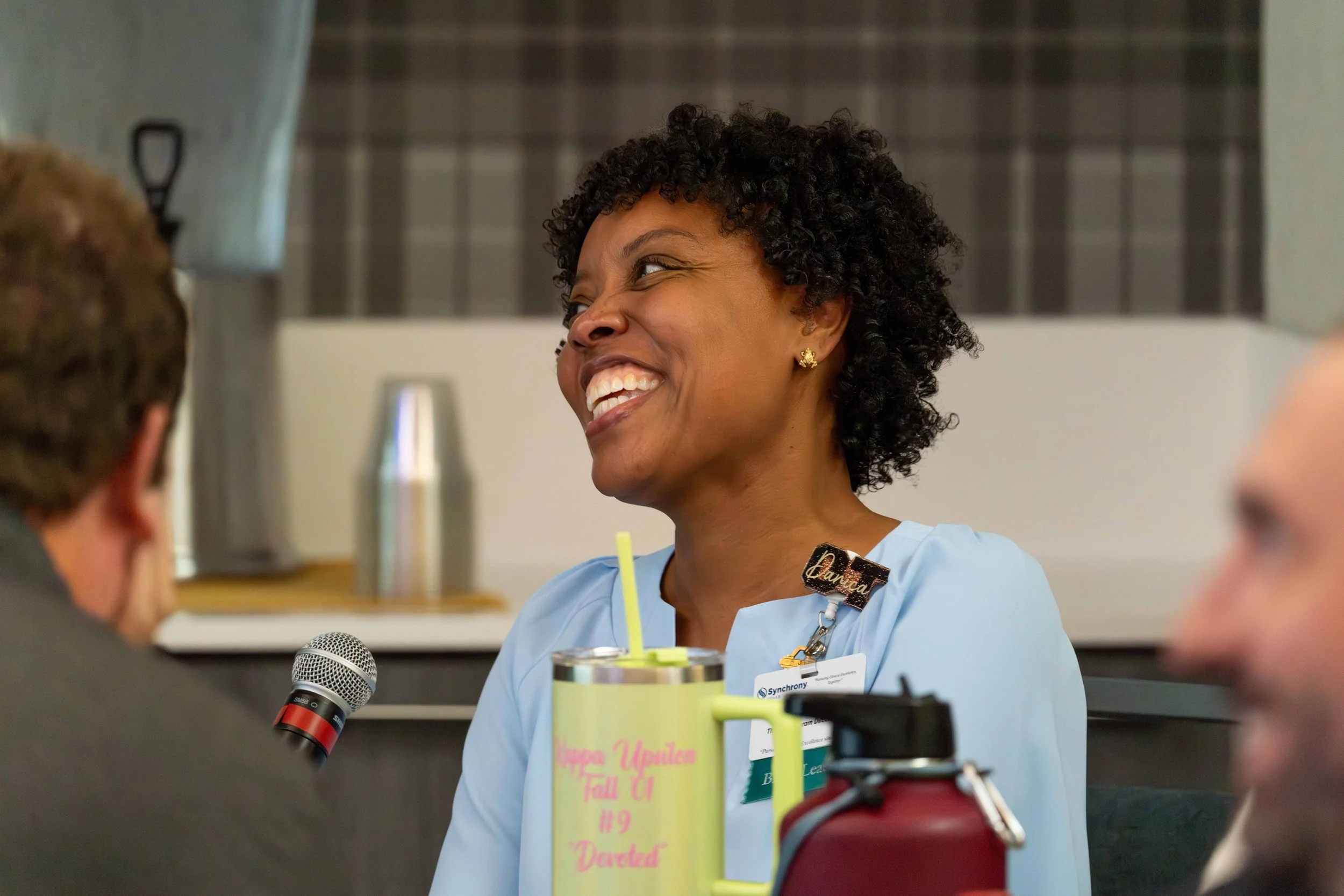 A smiling woman with curly hair wearing a light blue nurse uniform and name badge, talking to individuals in a meeting or discussion, with a microphone and water bottle on the table.