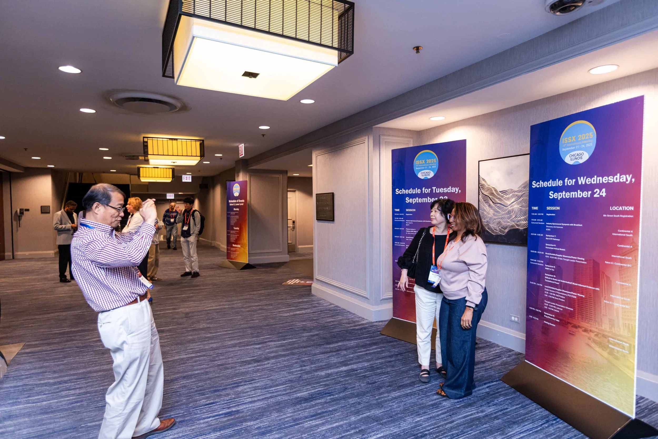 Conference lobby with people taking pictures and talking near large informational posters about schedules, with a man in checkered shirt taking a photo of two women side by side.