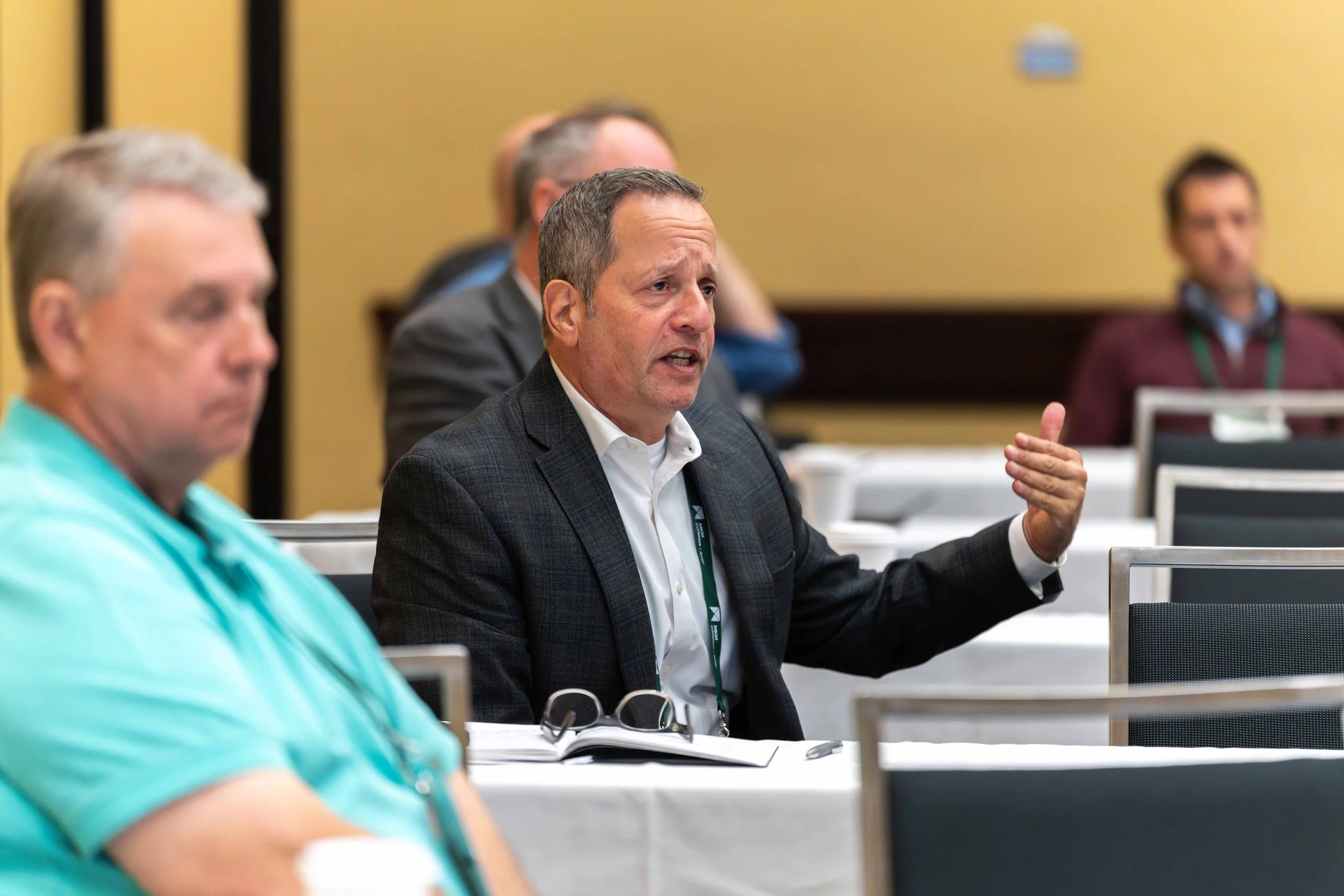 A man in a dark checkered blazer and white shirt speaking during a conference, with others seated around him in a meeting room.