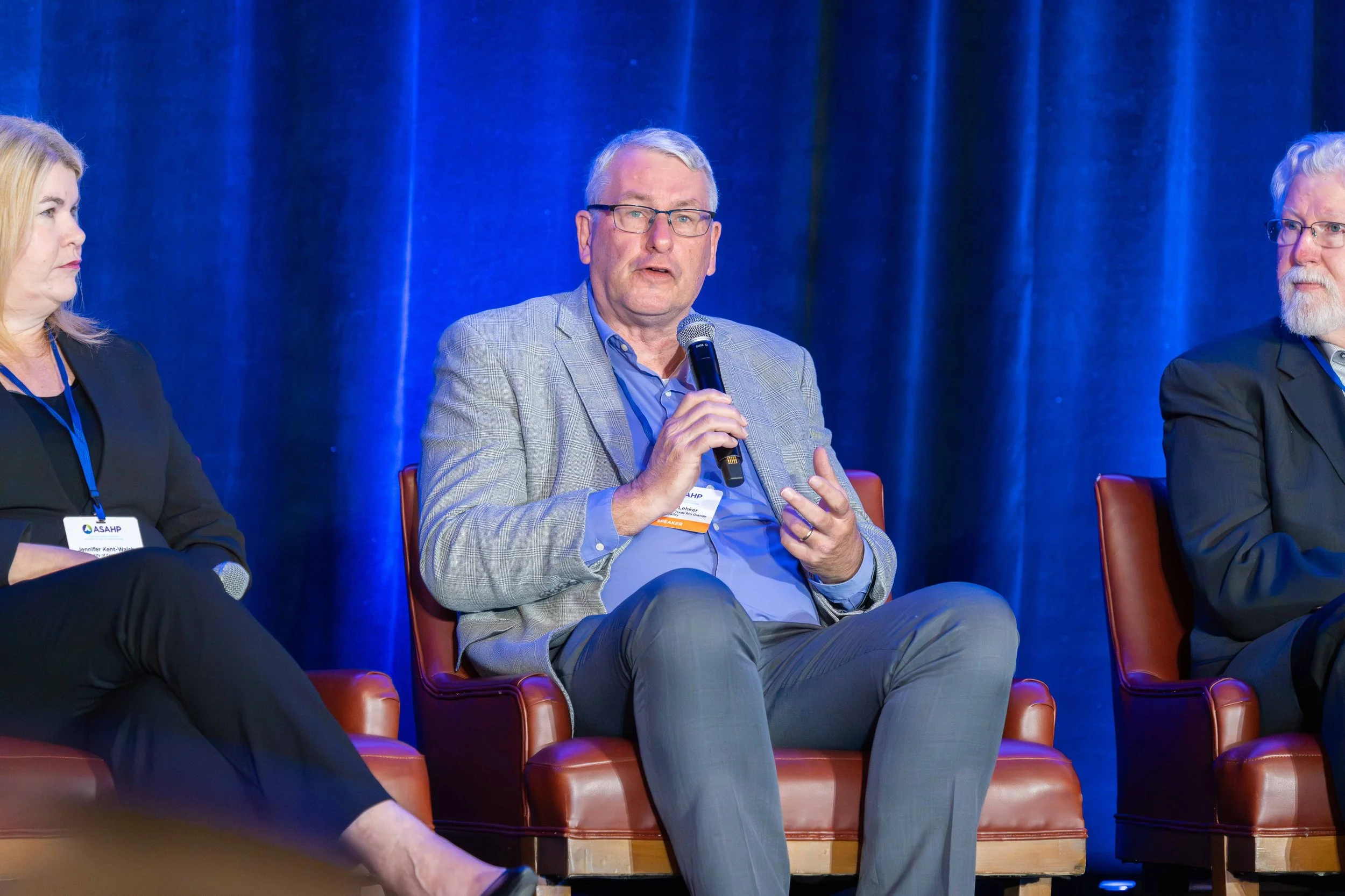 A man in a gray plaid suit and glasses speaking into a microphone during a panel discussion. He is sitting on a brown leather chair with a blue curtain background.