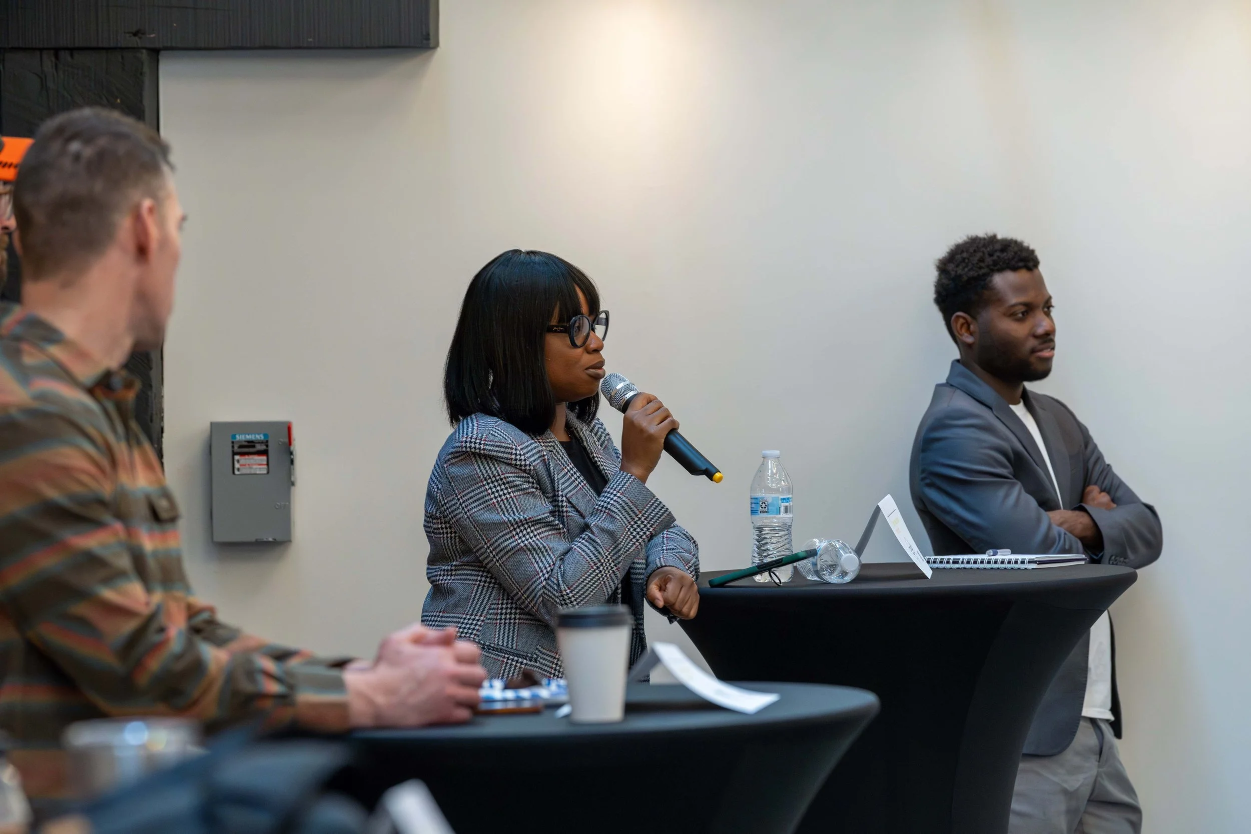 A woman with glasses speaking into a microphone at a panel discussion. She is seated at a table with a water bottle, a paper cup, and notes. Two men are seated at the same table, one in a patterned shirt, the other in a gray suit, listening attentive