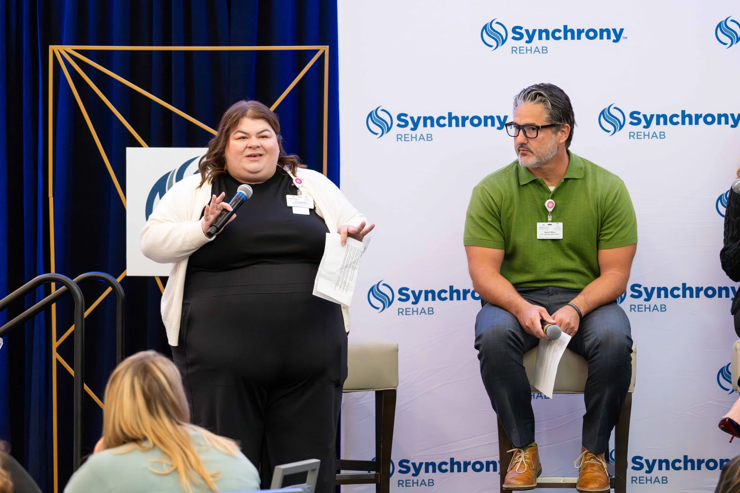A woman holding a microphone and a sheet of paper standing next to a seated man holding a microphone during a panel discussion at a Synchronic Rehab event. The background features a white wall with the Synchronic Rehab logo repeated and dark blue cur