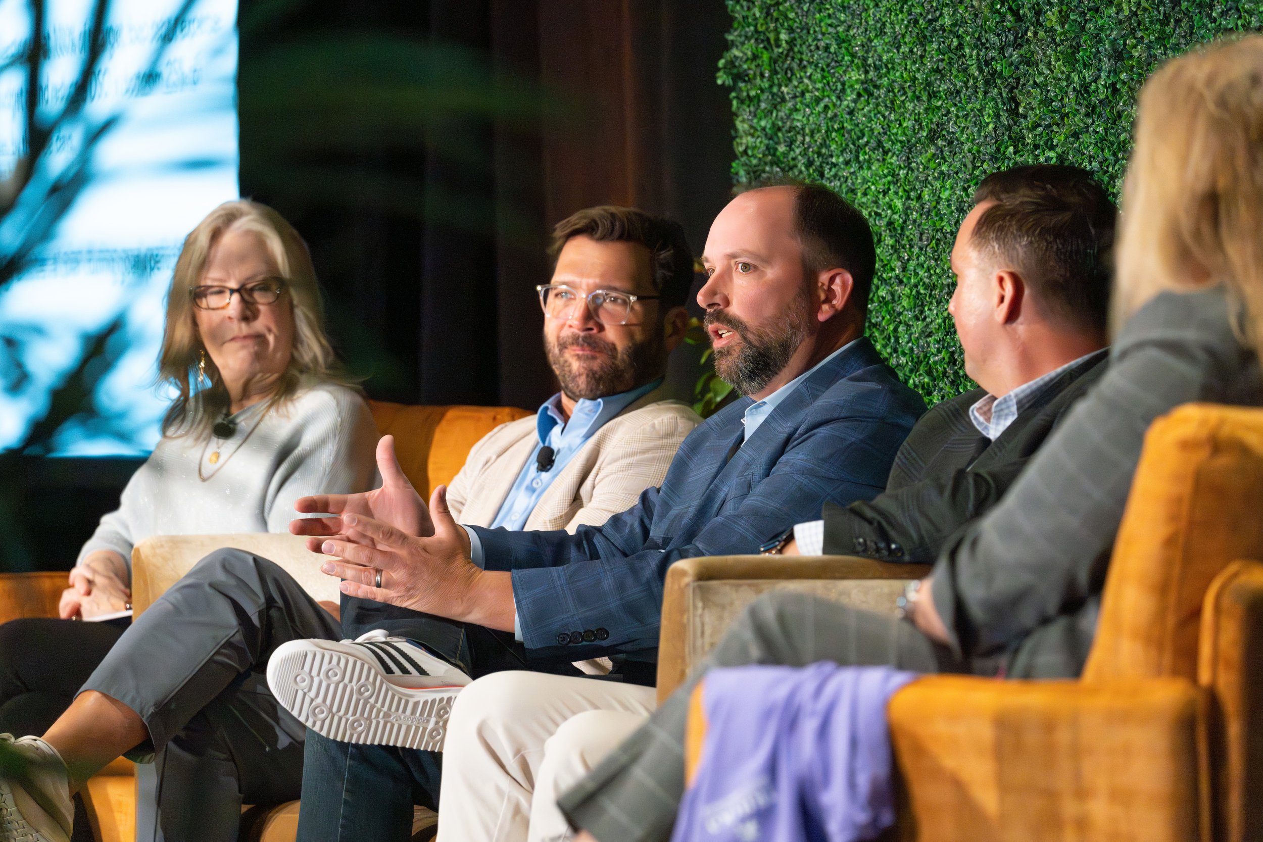 Five individuals seated on a stage engaged in a panel discussion, with a woman on the left, a man in the middle speaking, and four people in suits listening.