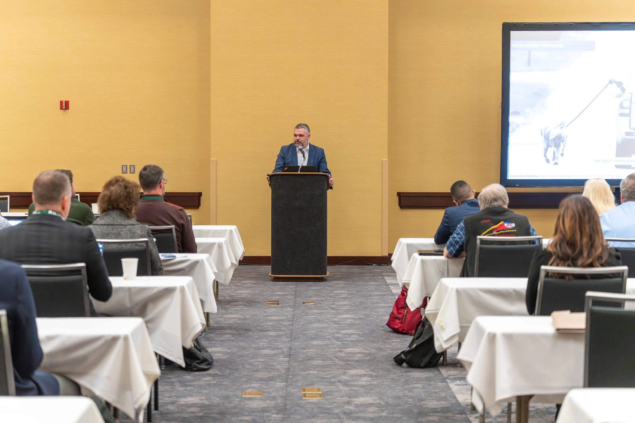 A man in a blue blazer giving a presentation at a conference, standing behind a podium. Attendees seated at white-covered tables listen, with a large screen displaying a slide in the background.