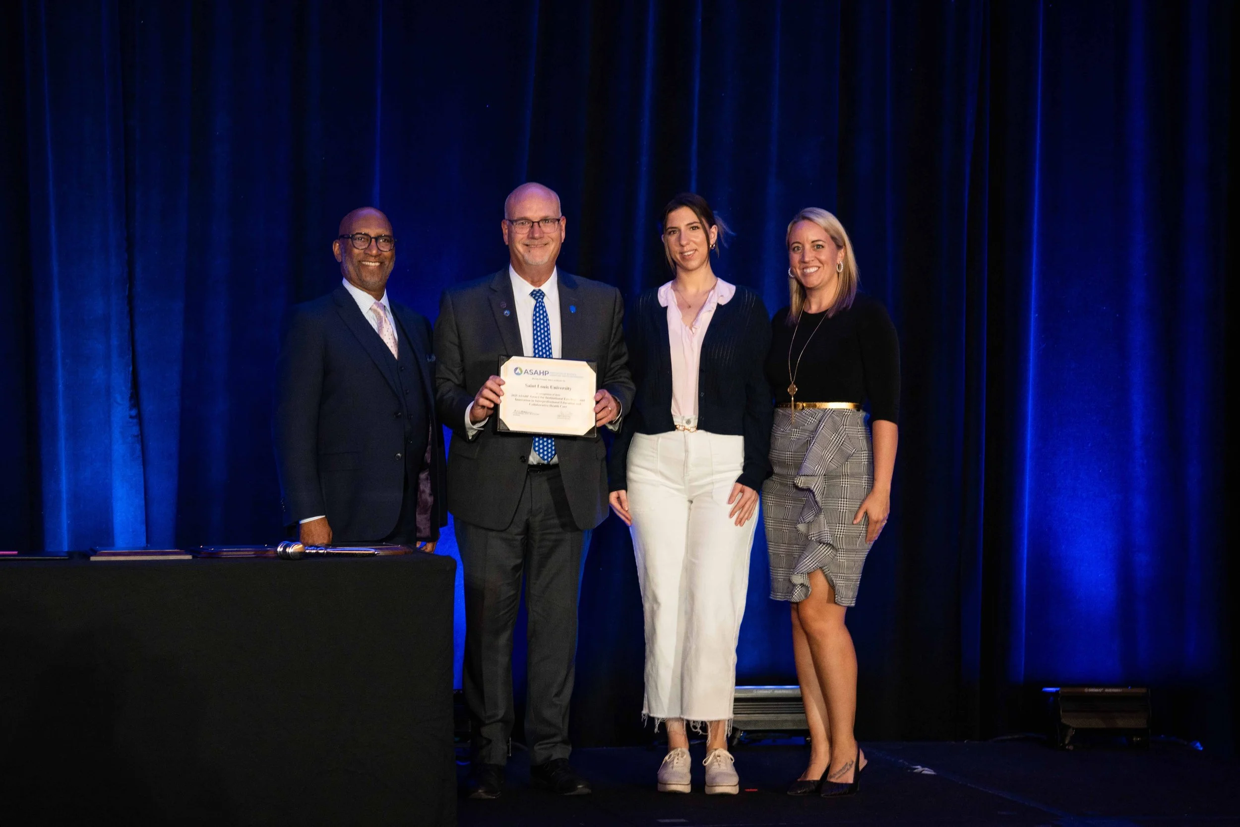 Group of four people standing on stage during an award ceremony, with dark curtains in the background. The man second from the left is holding a certificate, smiling.