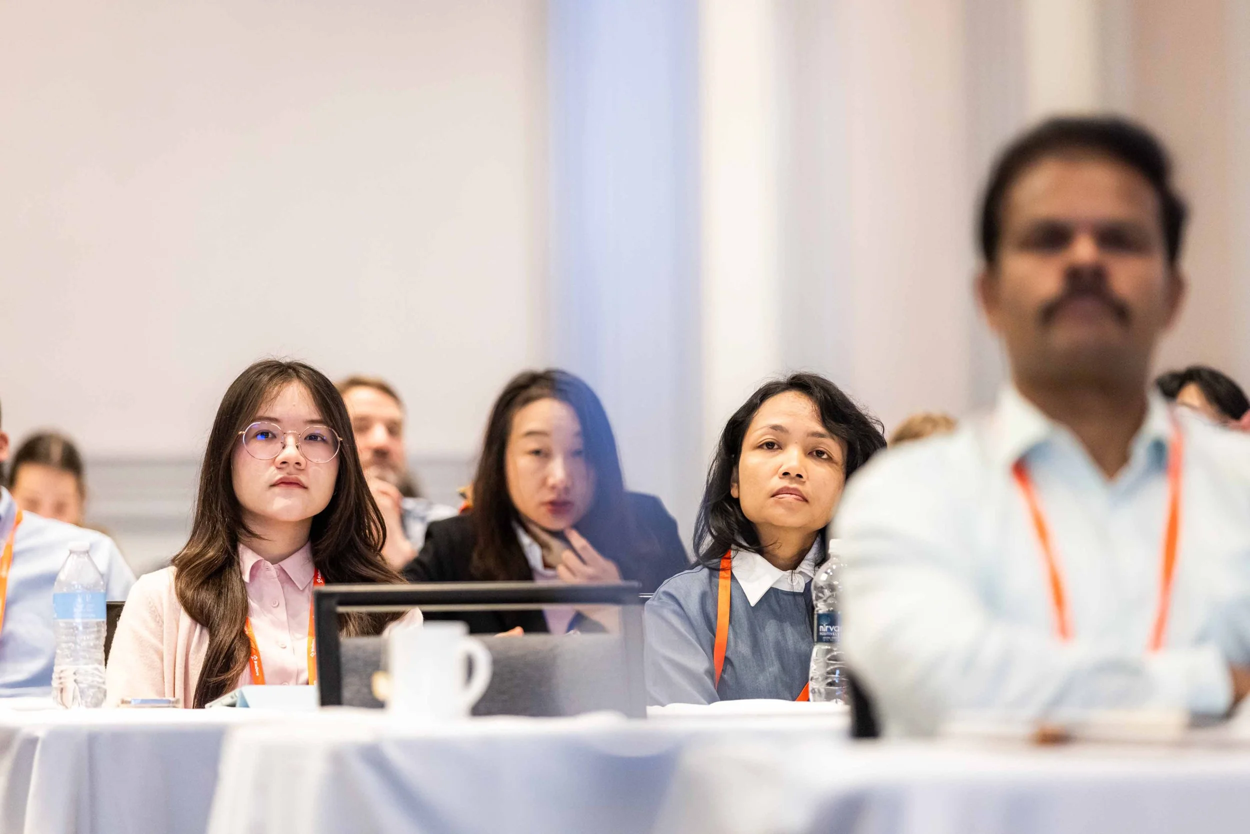 Attendees at a conference sitting at tables, listening attentively, with water bottles and notebooks in front of them.