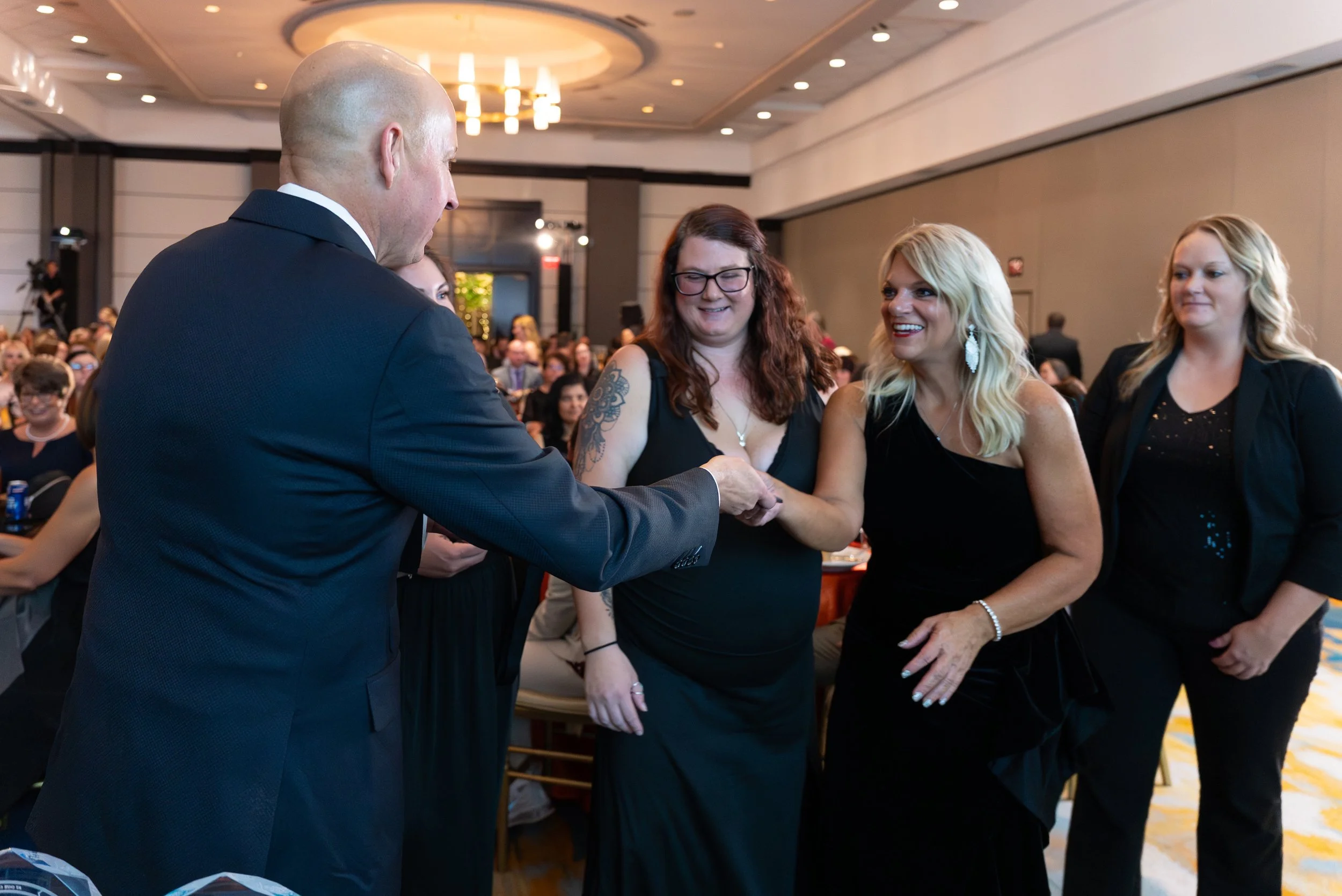 A man in a suit shaking hands with a smiling woman in a black dress at an indoor event, with other women and a large audience in the background.