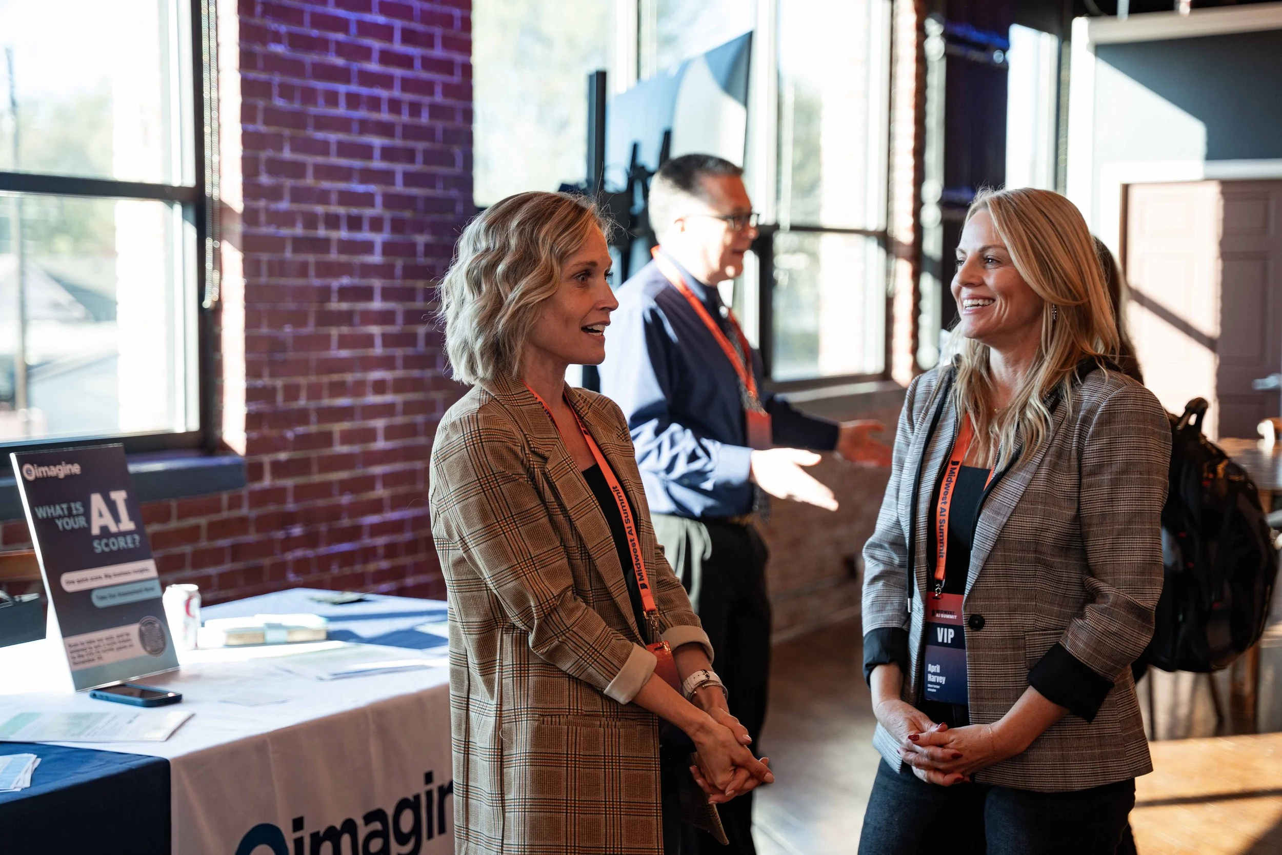 Two women and one man engaging in conversation at a professional event. The women are smiling and wearing name tags, and the man is standing in the background near a window with brick walls.