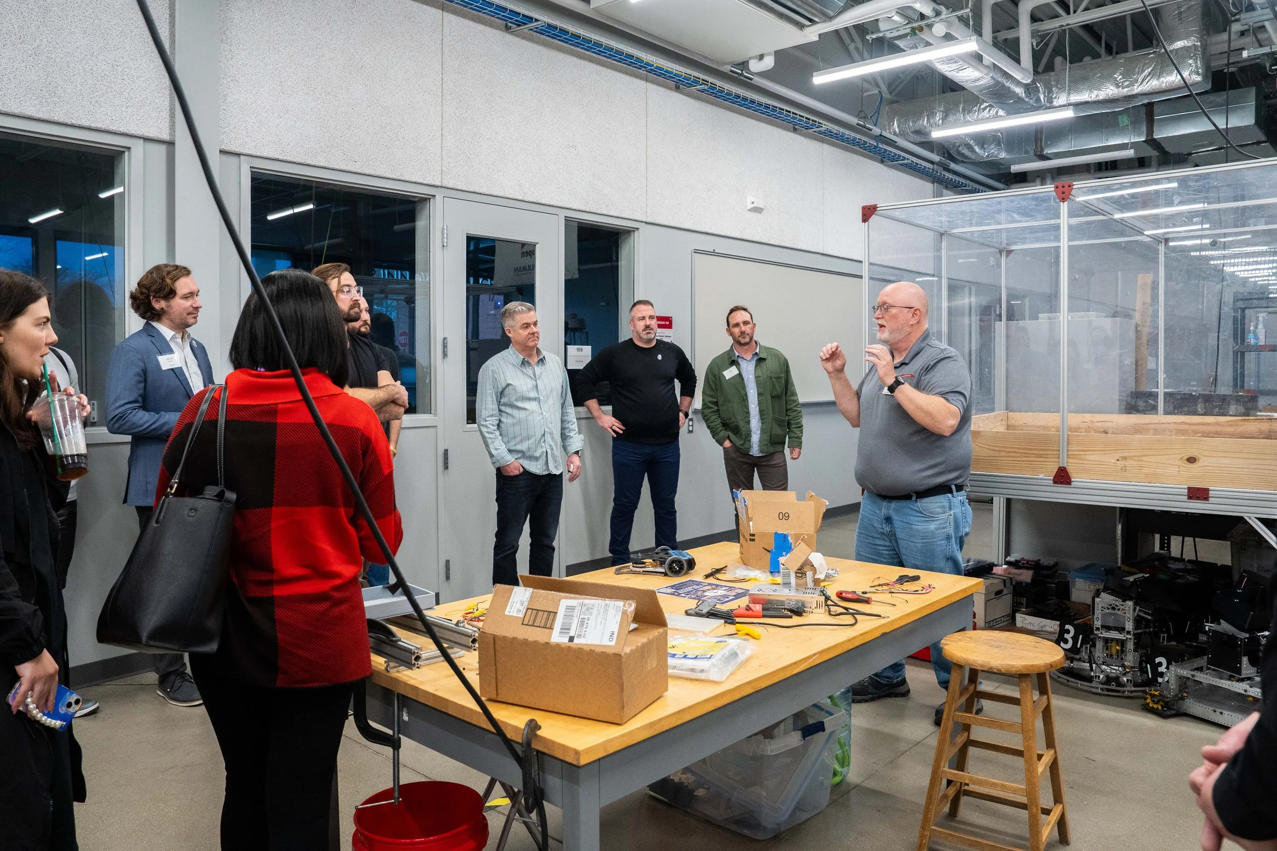 A group of people listening to a man giving a presentation in a workshop or classroom setting with various tools and equipment on a work table.