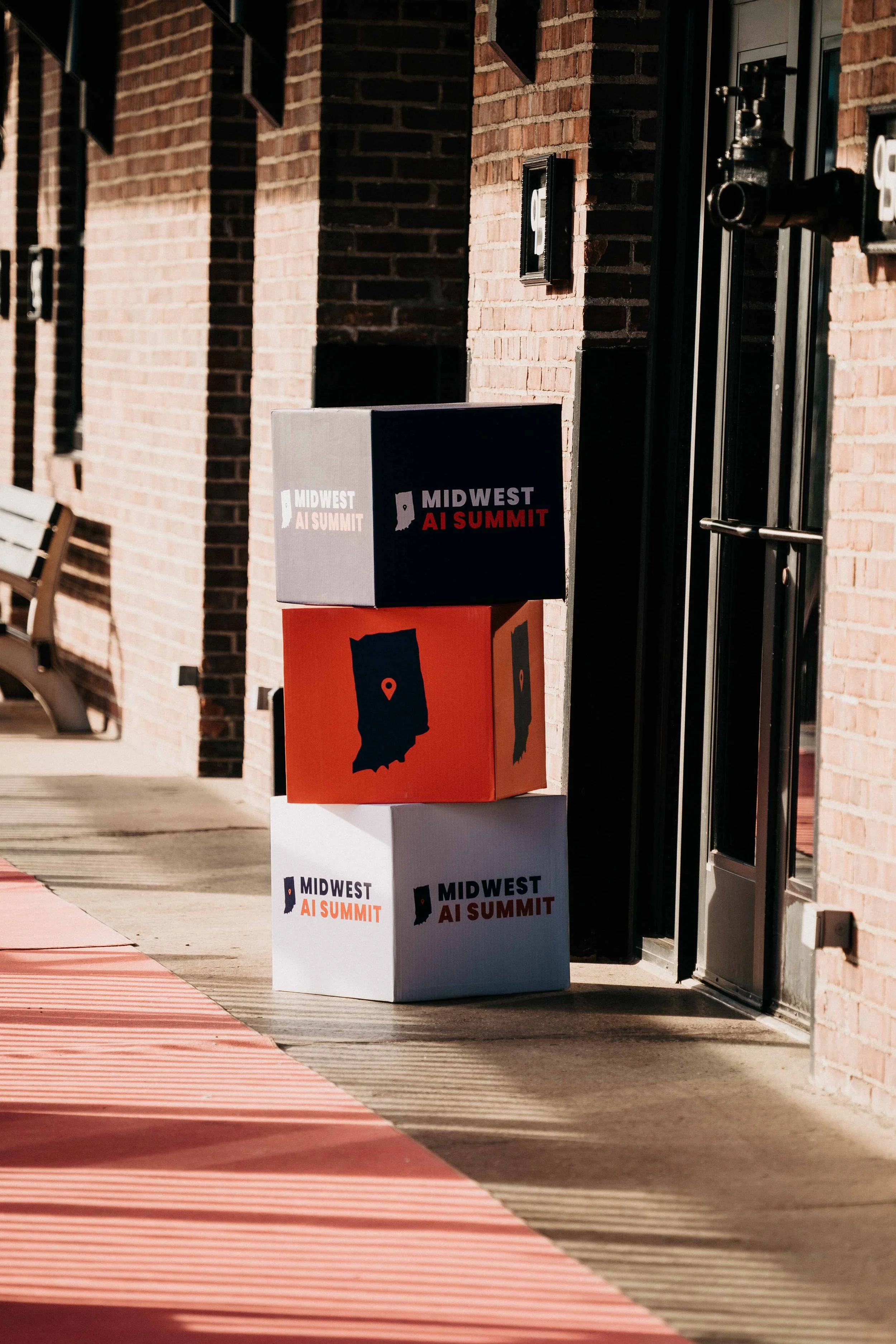 Three stacked boxes on a sidewalk outside a brick building, advertising the Midwest AI Summit, with logos and map of Indiana.