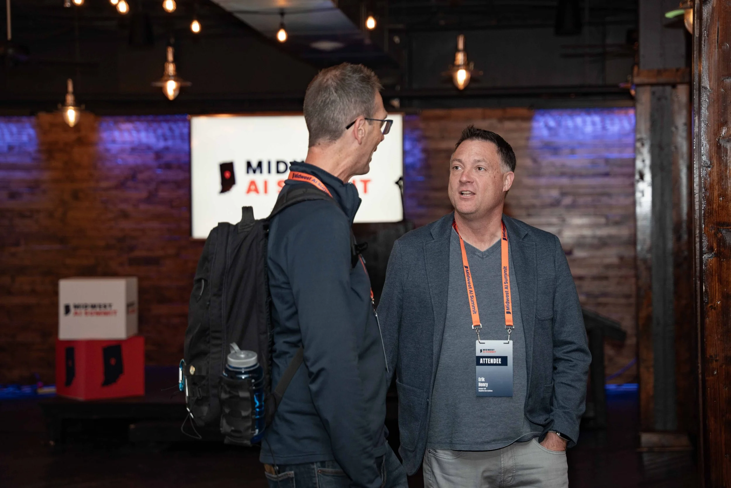 Two men talking at a conference or event, with one wearing glasses, a backpack, and a lanyard, and the other with a conference badge that says 'ATTENDEE'.