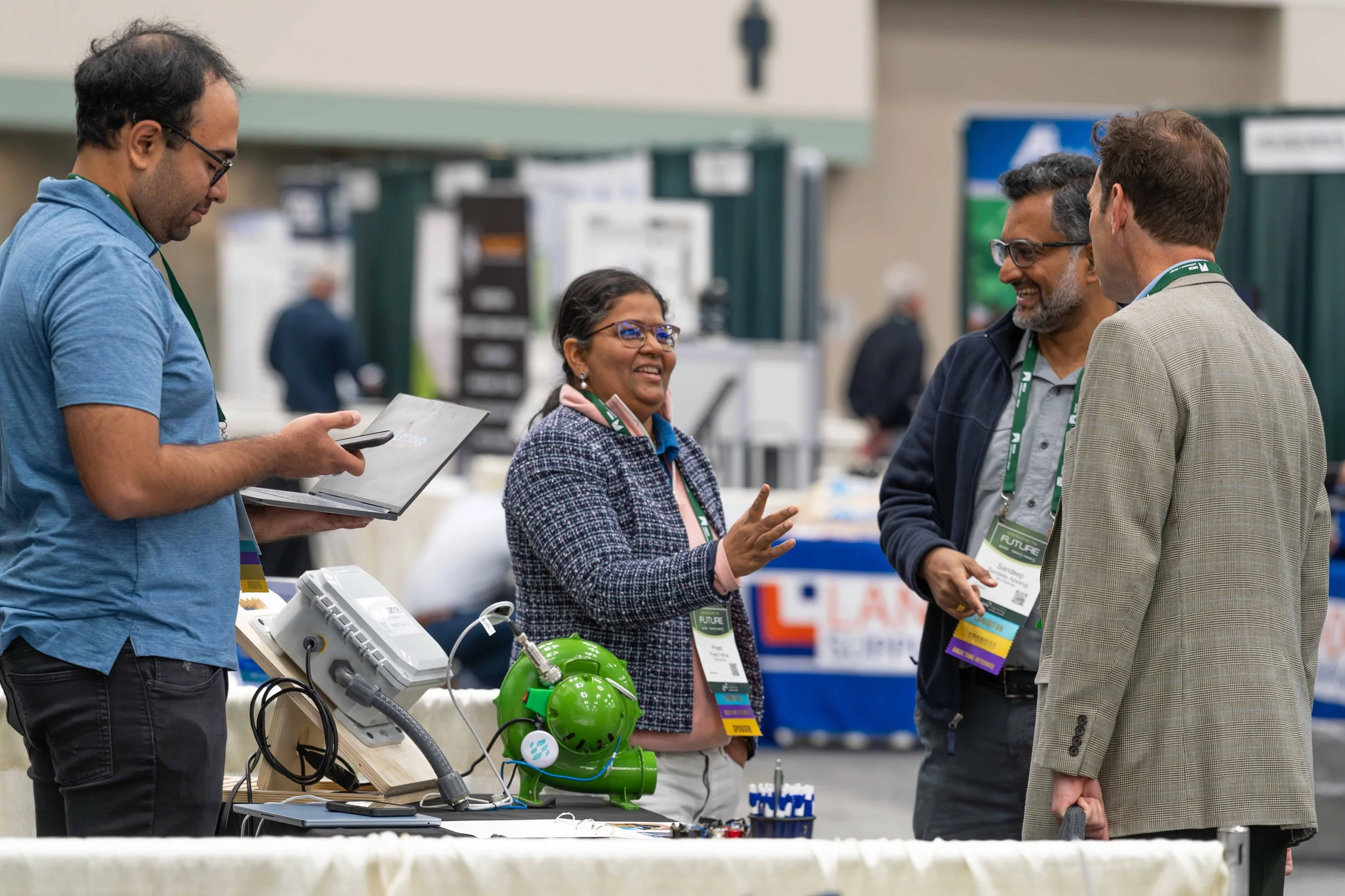 Four people are engaged in conversation at a conference or exhibition, with a table displaying electronic equipment, including a green device, some wires, and a vintage telephone.