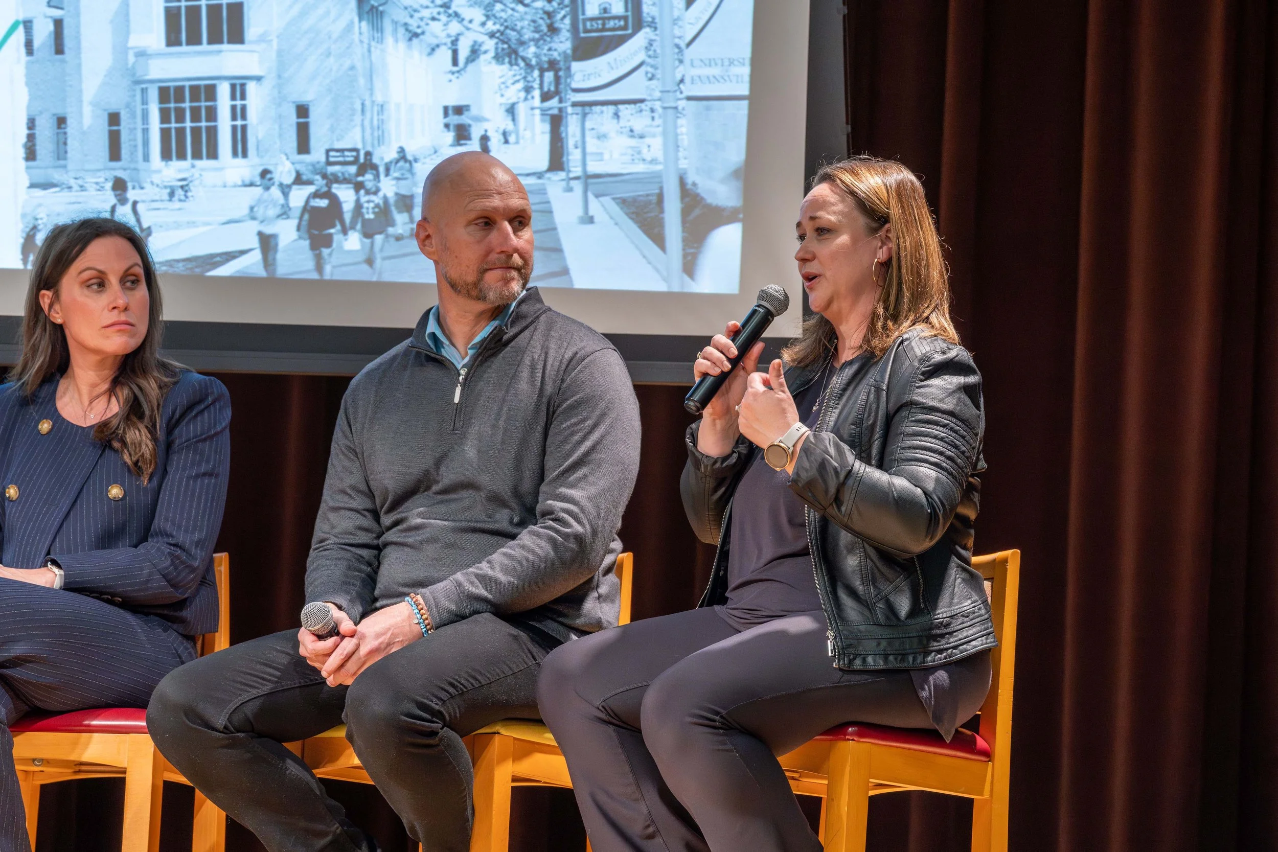 Three people sitting on stage during a panel discussion, with two holding microphones and one speaking, and a large screen behind displaying a black-and-white photo of people walking on a university campus.