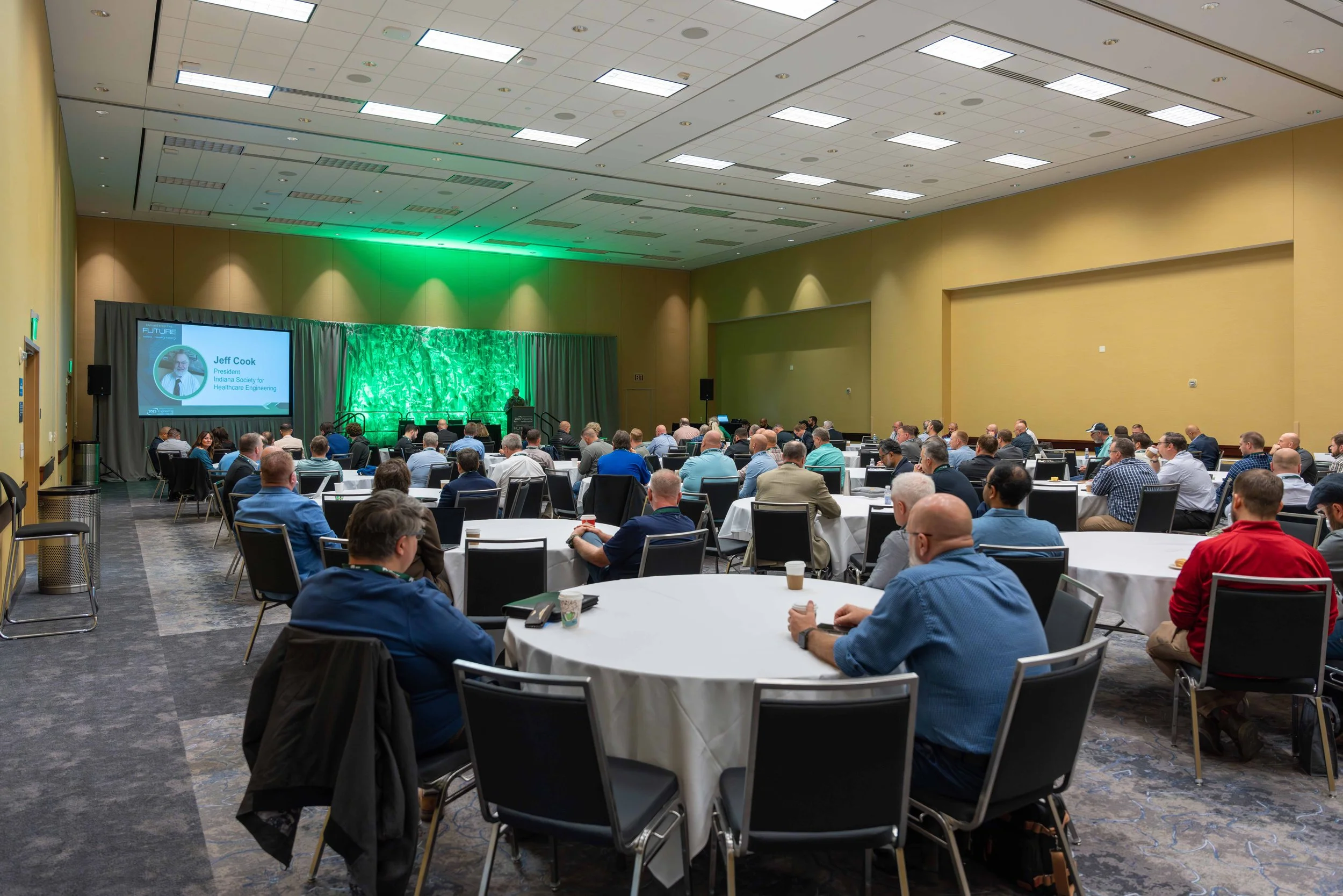 Large conference room filled with attendees seated at round tables, facing a stage with a screen displaying a speaker named Jeff Cook, President of Indiana Society for Healthcare Engineering, with green lighting behind the stage.