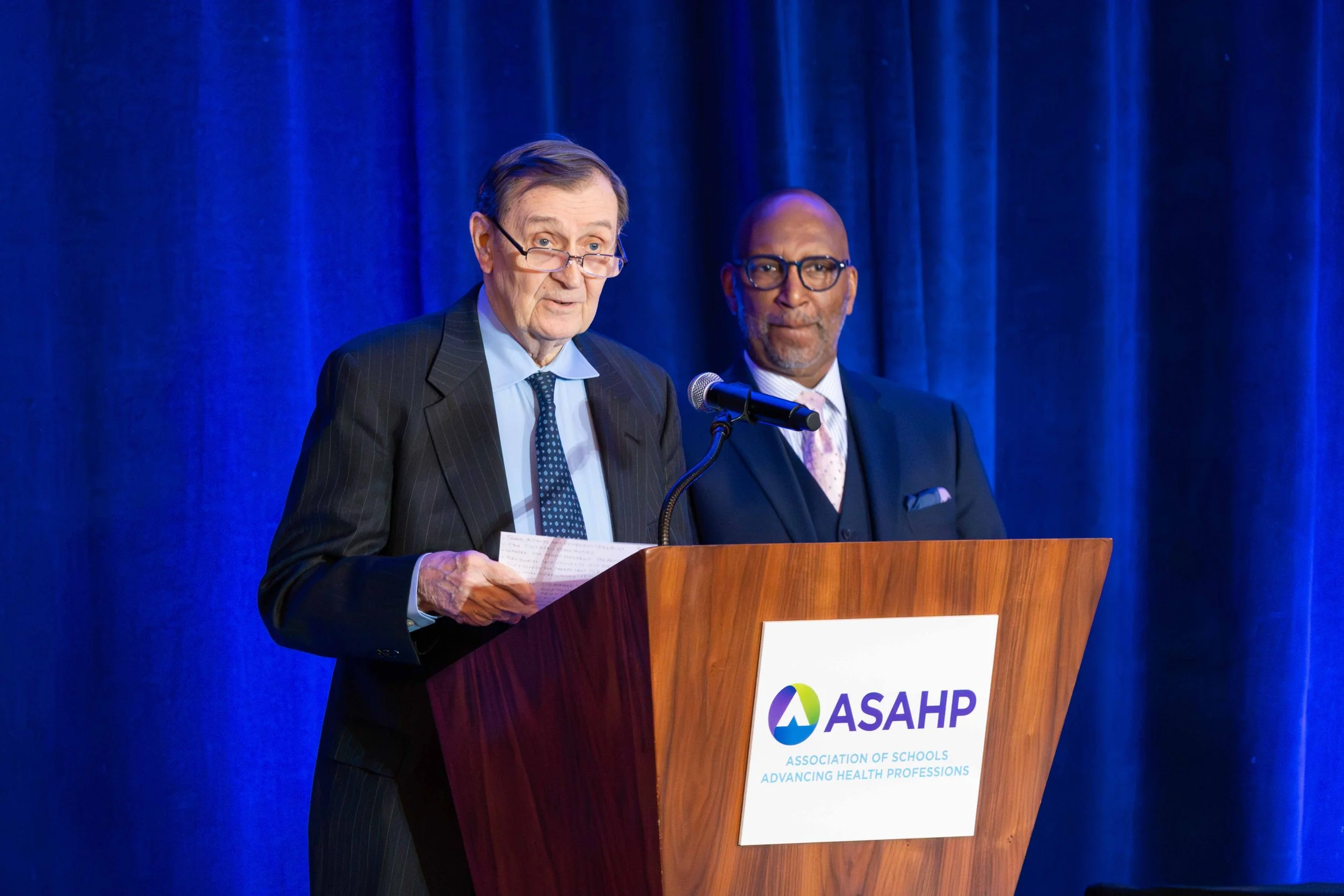 Two men standing at a wooden podium with a blue curtain backdrop, speaking at an event for the Association of Schools Advancing Health Professions (ASAHP).