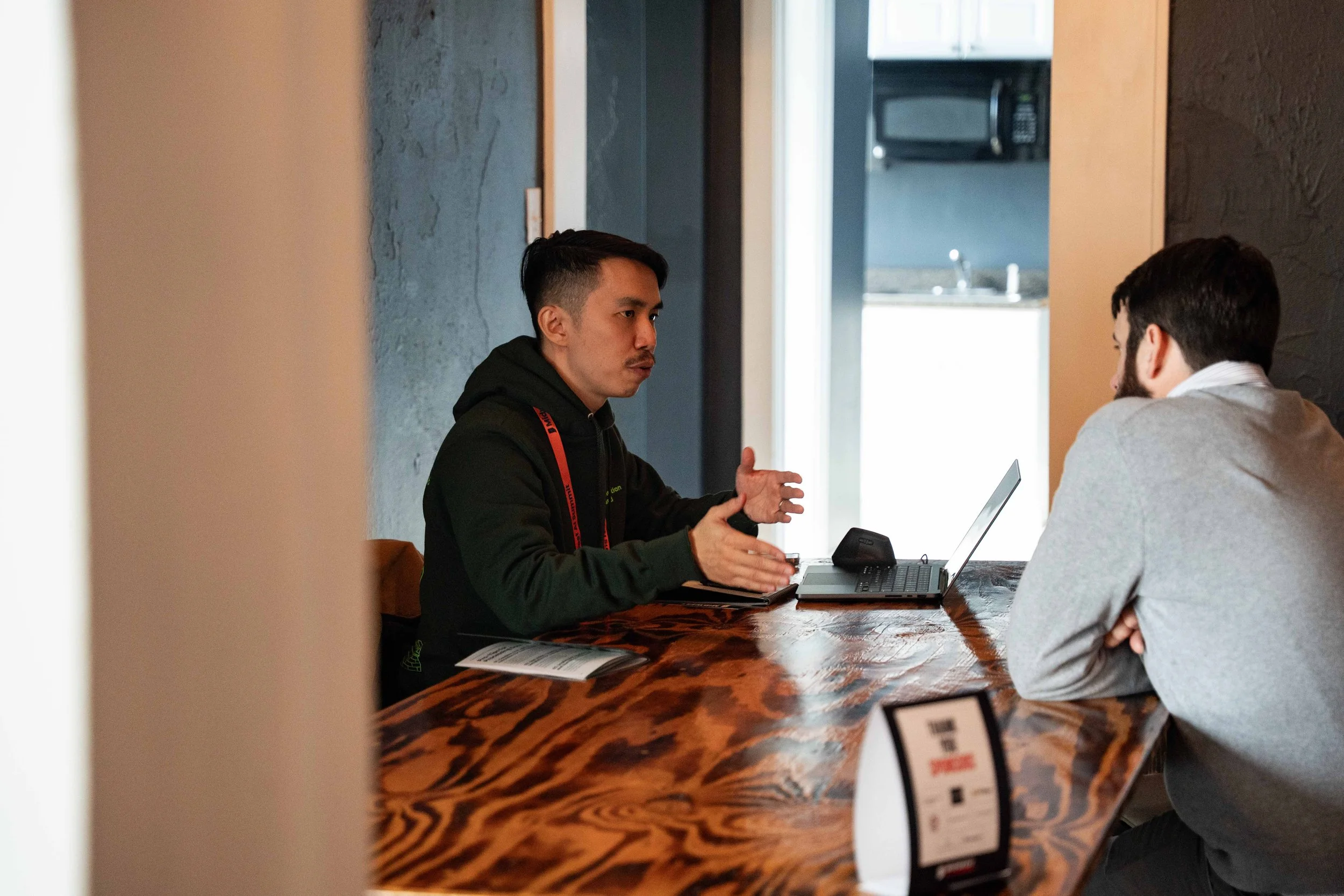 Two men having a discussion at a wooden table in a modern setting, with a laptop and documents.