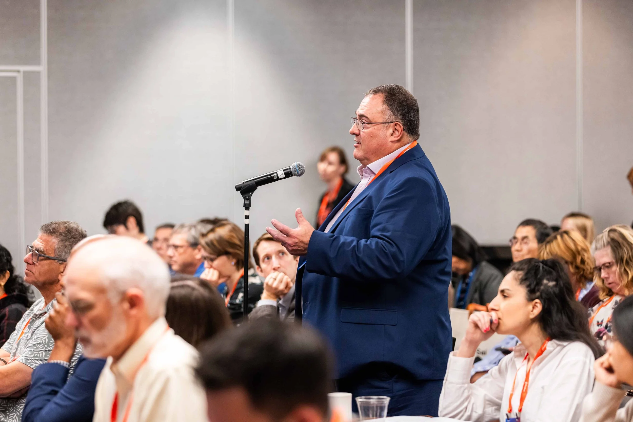 Man in blue suit speaking into a microphone at a conference, audience members listening and taking notes.