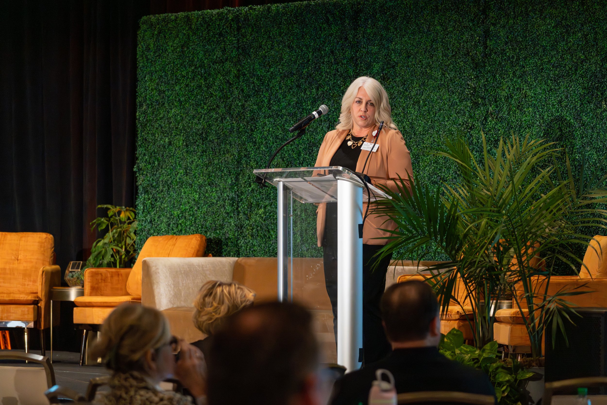 A woman with blonde hair wearing a tan blazer speaking at a podium on stage with a green leafy background, while audience members listen, some taking photos or notes, with orange chairs and green plants around.