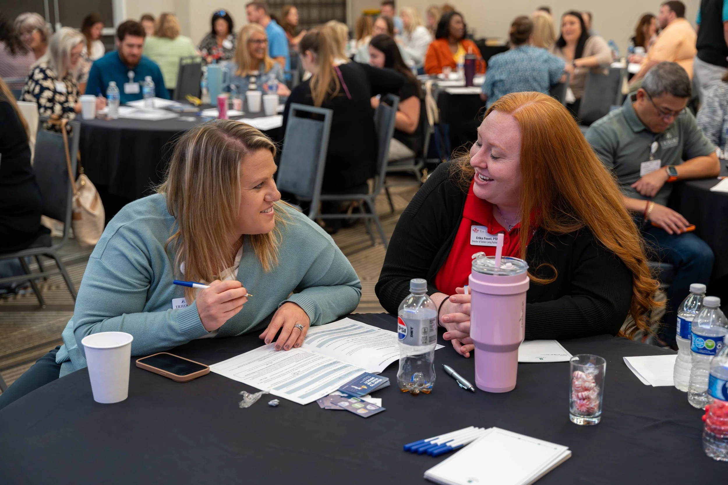 Two women are sitting at a conference table, engaging in conversation, surrounded by other conference attendees in a large room.