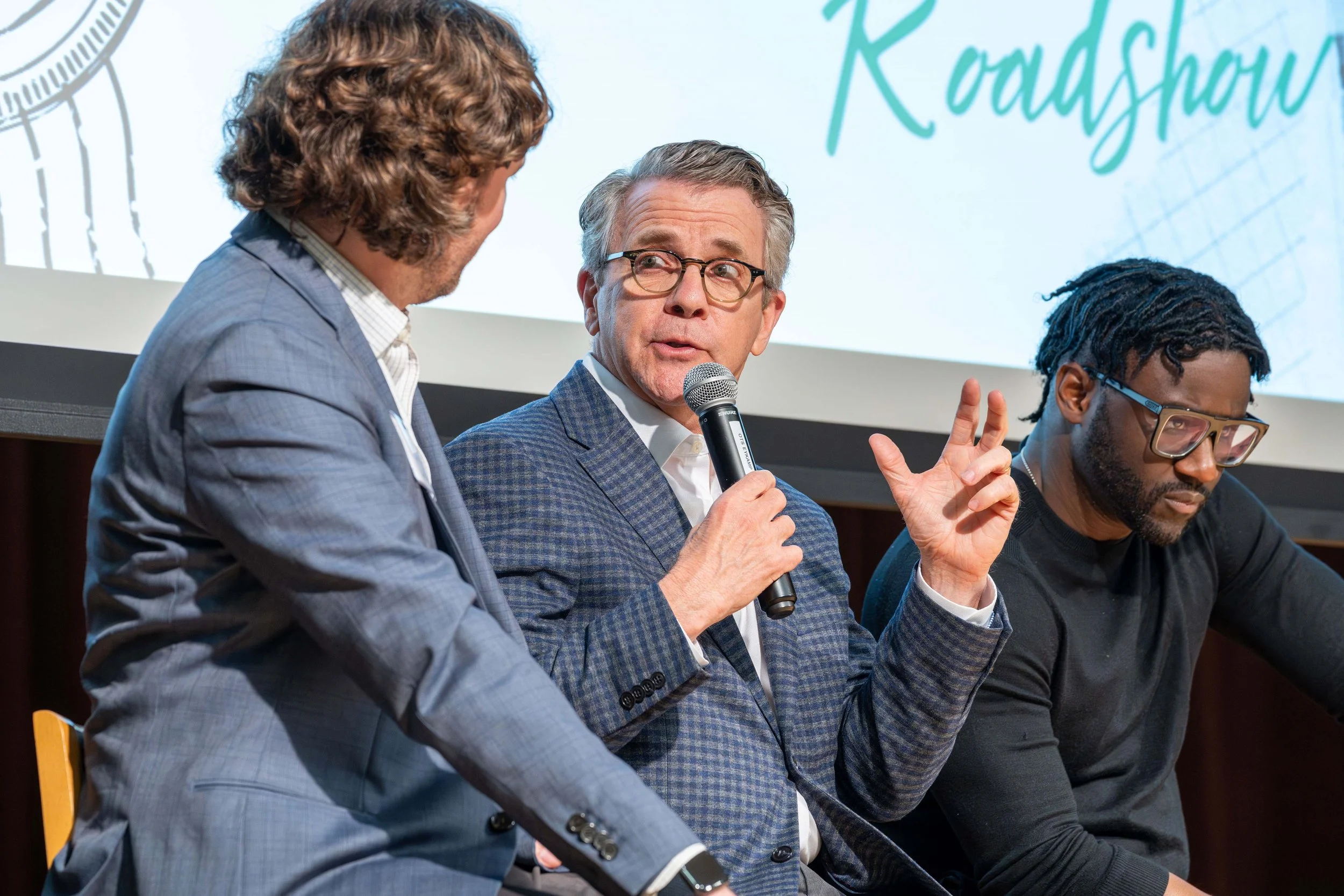Three men sitting at a panel discussion, with one man speaking into a microphone, in front of a screen with the word 'Roadshow' on it.
