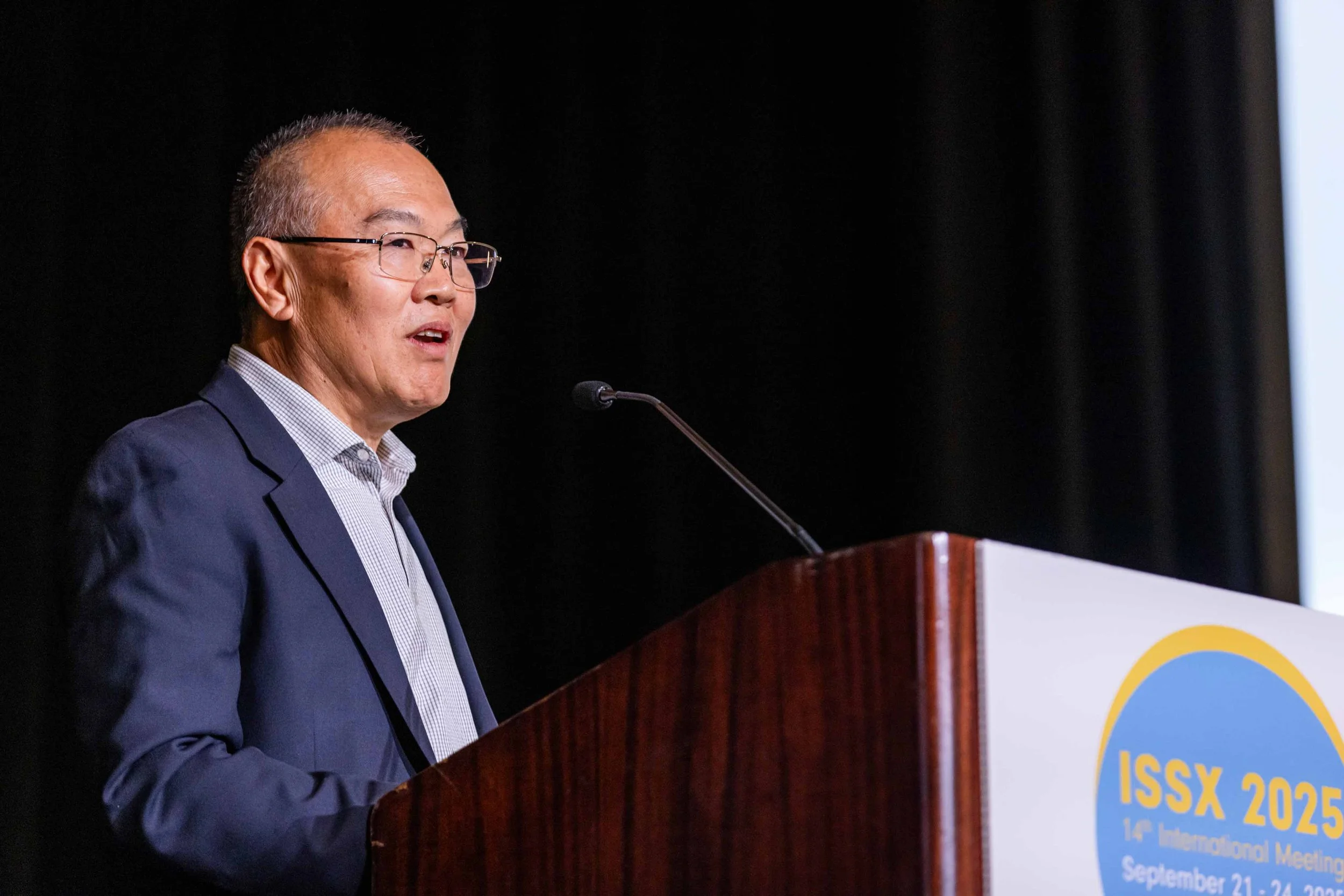 A man in a suit speaking at a podium with a sign that reads 'ISSX 2025, 14th International Meeting, September 21-24, 2025'.