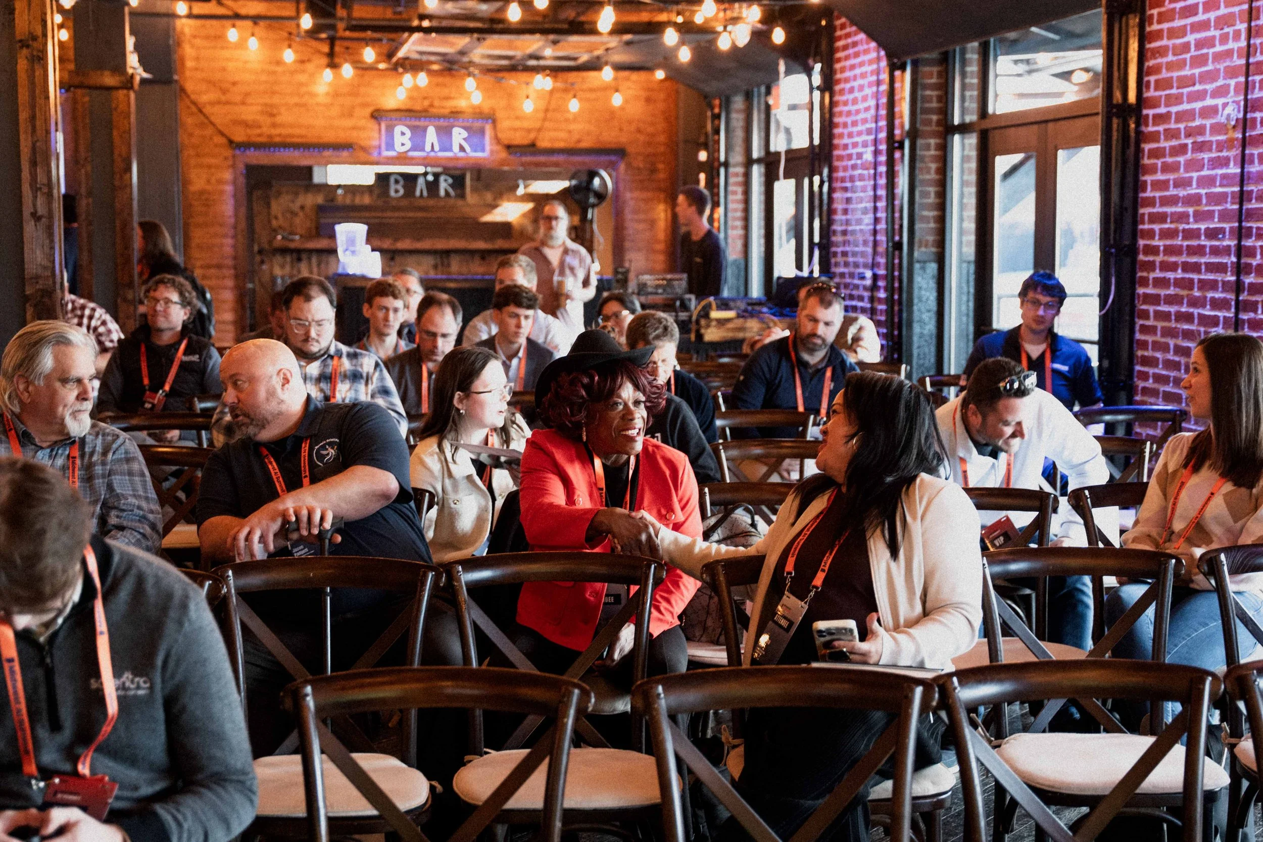 People sitting in a conference room, engaging in conversations and listening to a speaker. The room has wooden walls, brick accents, large windows, and string lights. Two women in the front are shaking hands.