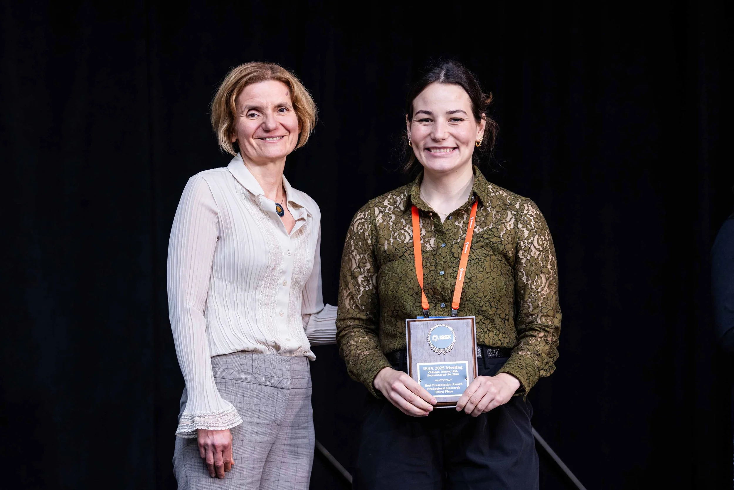 Two women standing together, smiling, at an award ceremony. The woman on the right is holding a plaque and wearing a conference badge.