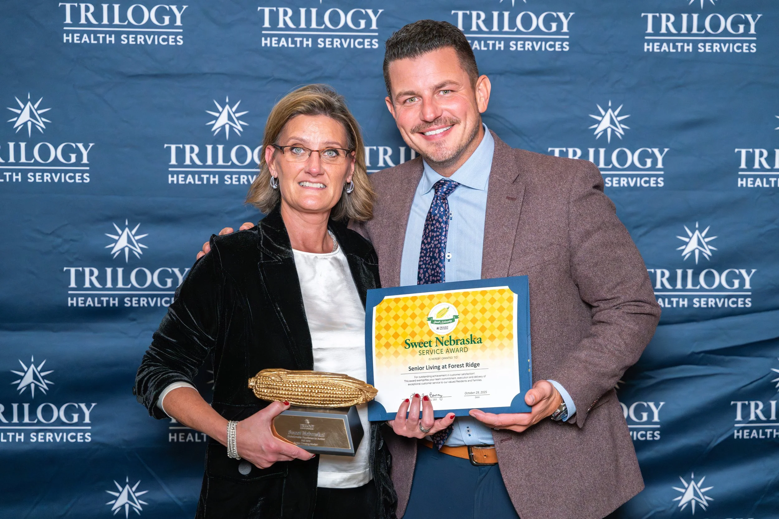 Two people standing together at an awards ceremony, holding a certificate and a trophy. The backdrop displays 'TRILOGY HEALTH SERVICES'.